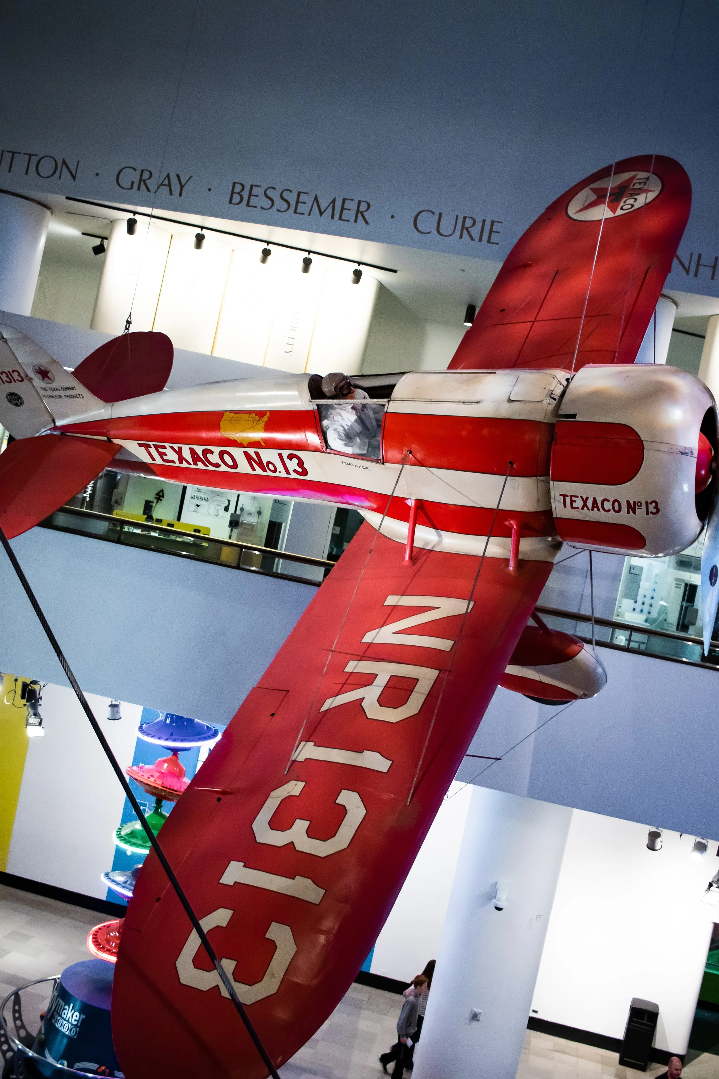 A red and white race plane with the text "Texaco No.13" on its side, displayed indoors, with visitors walking in the background and various displays in a museum setting.