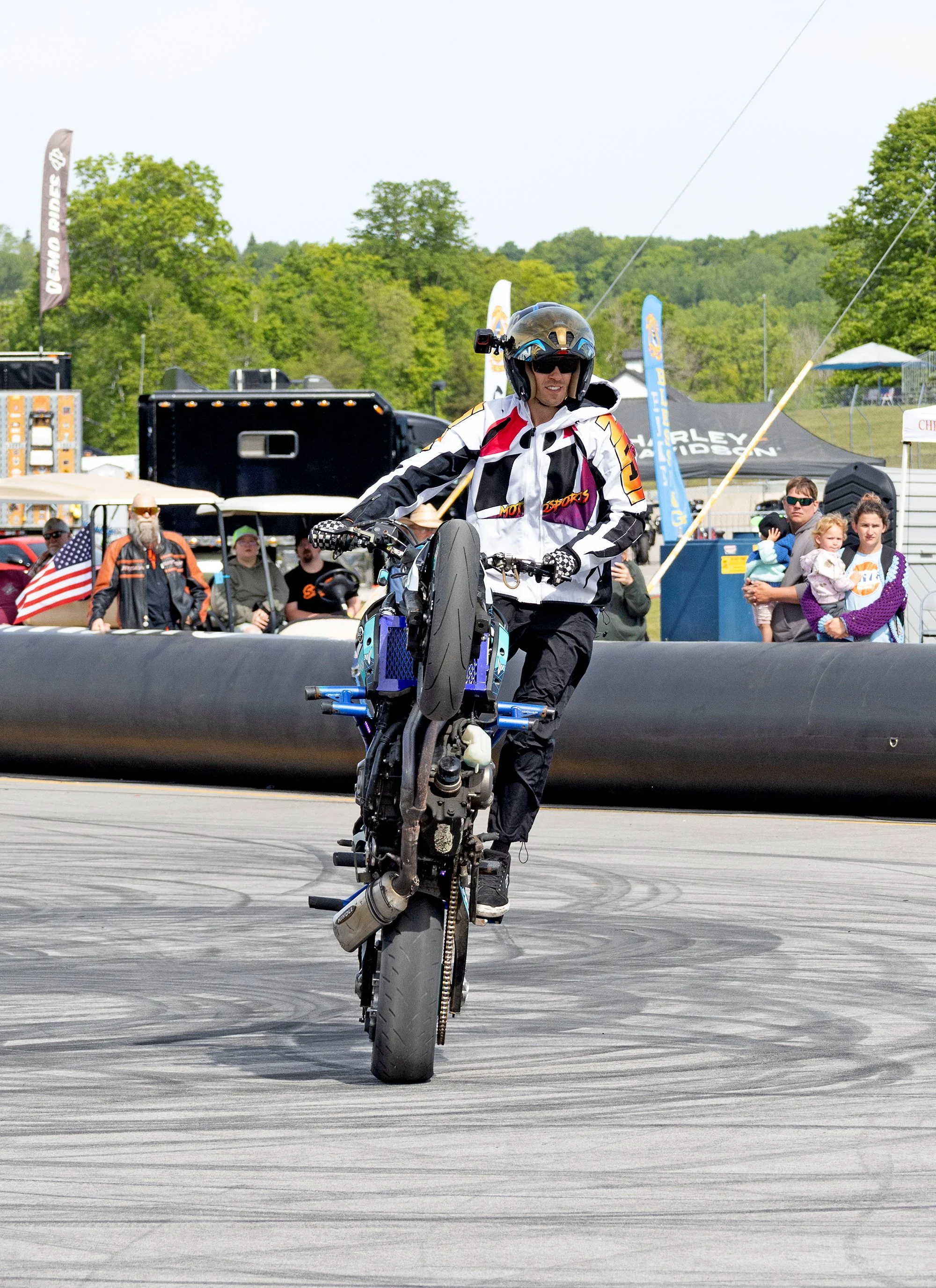 A person in motocross gear performing a stunt on a motorcycle at an outdoor event, with spectators and flags in the background.