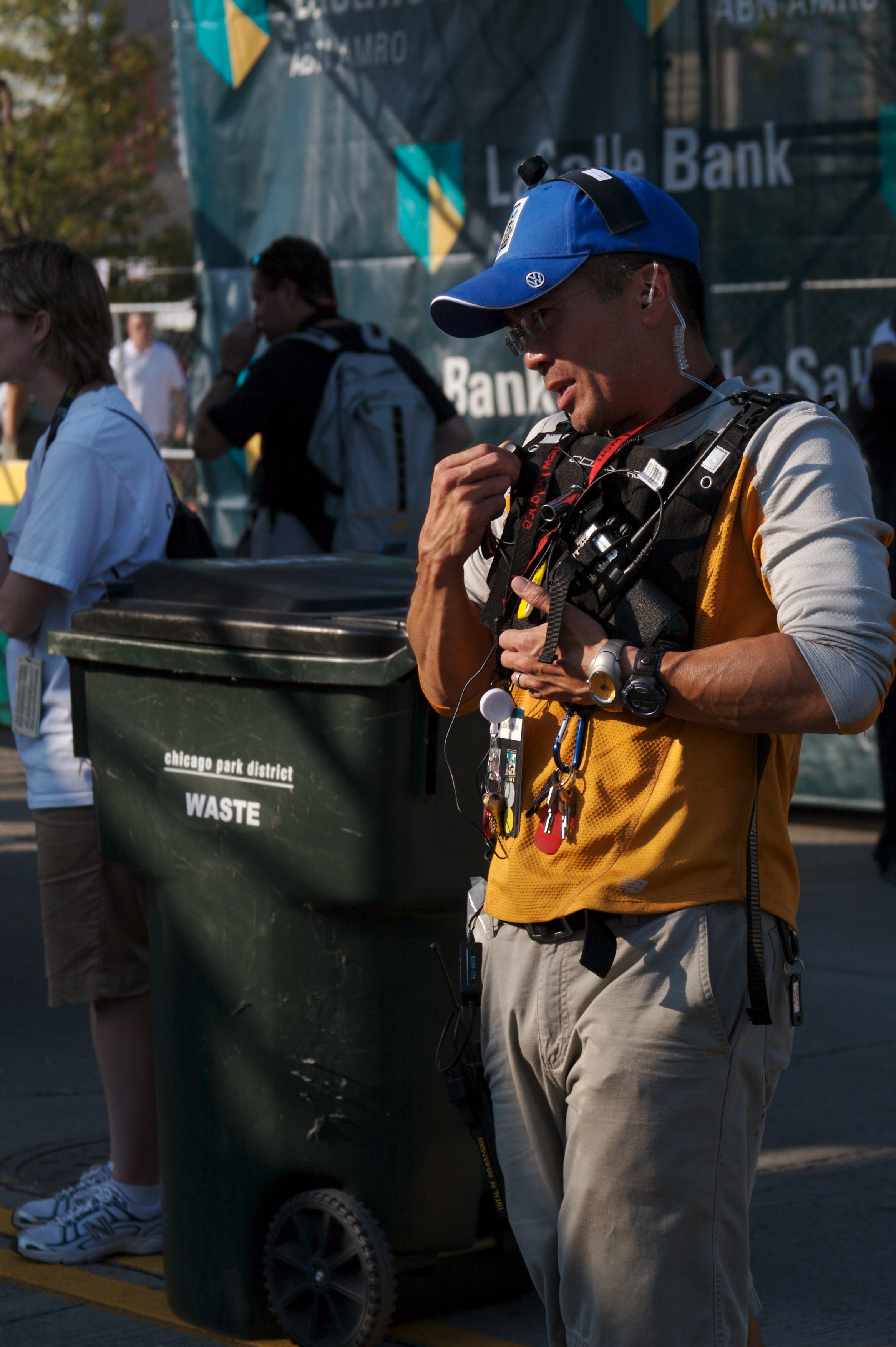 A man wearing a blue cap, glasses, and a yellow vest is standing next to a large green waste bin labeled 'chicago park district WASTE,' while holding a pen or similar object in his hand. In the background, there are other people, including a woman in