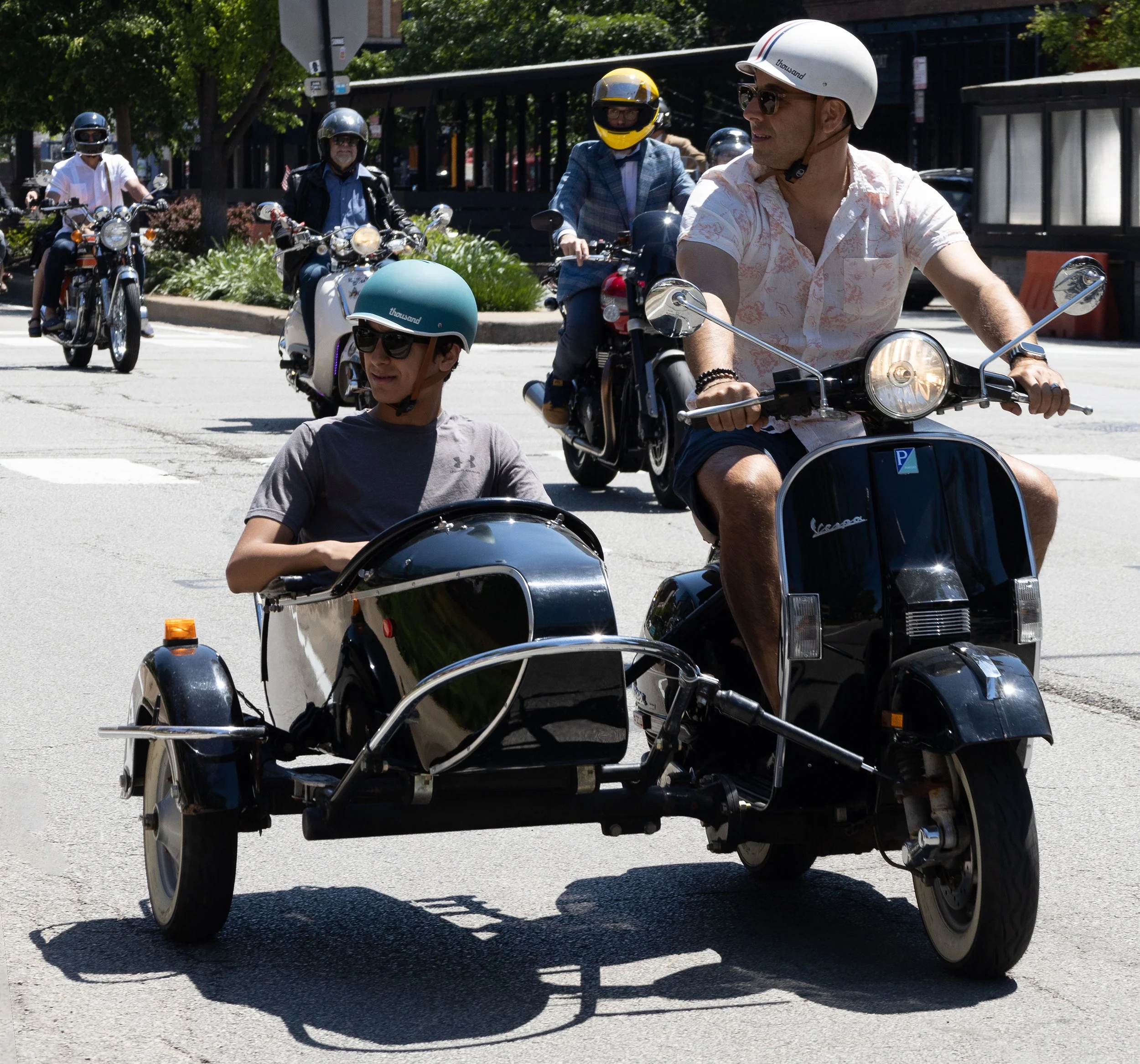 Multiple people riding motorcycles and a scooter on a city street, all wearing helmets, with some in sunglasses. The image captures a sunny day with trees and buildings in the background.