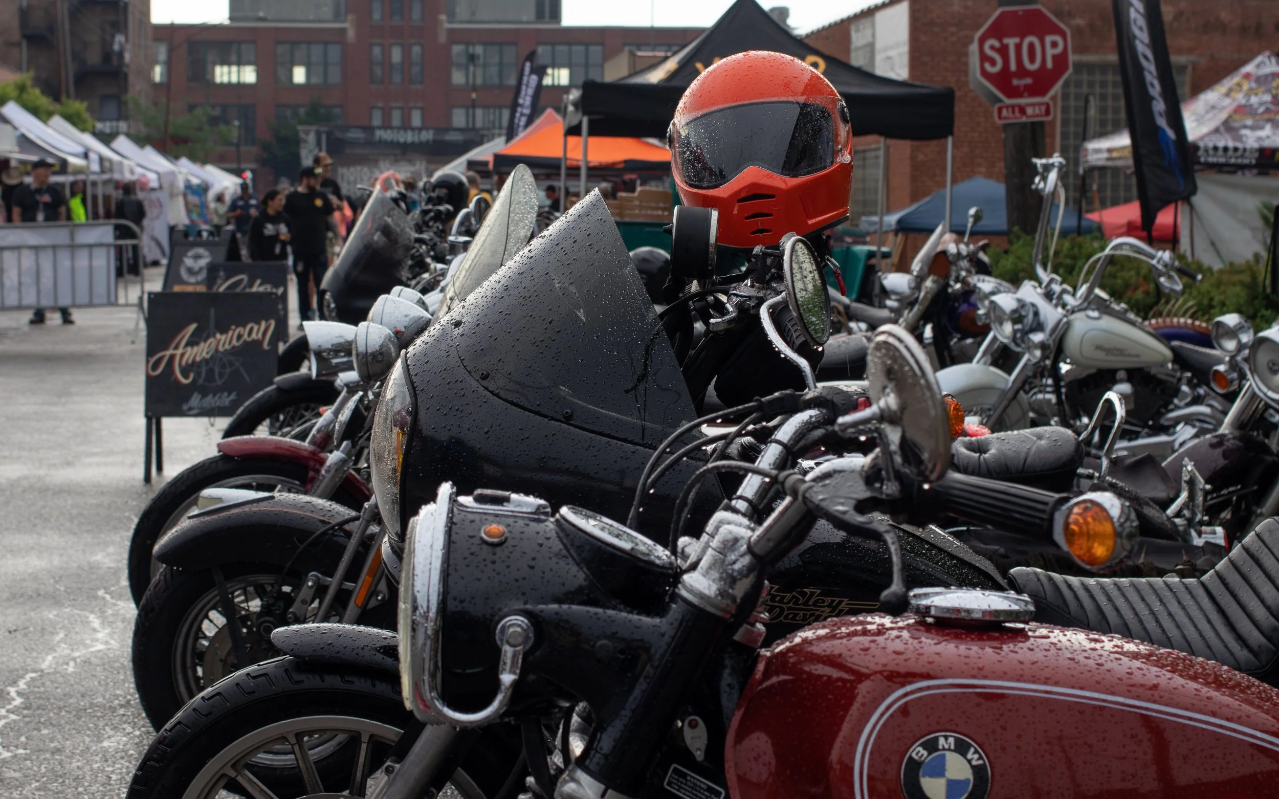 Multiple motorcycles parked at an outdoor event, with rain droplets visible on their surfaces and a red helmet resting on one of them.