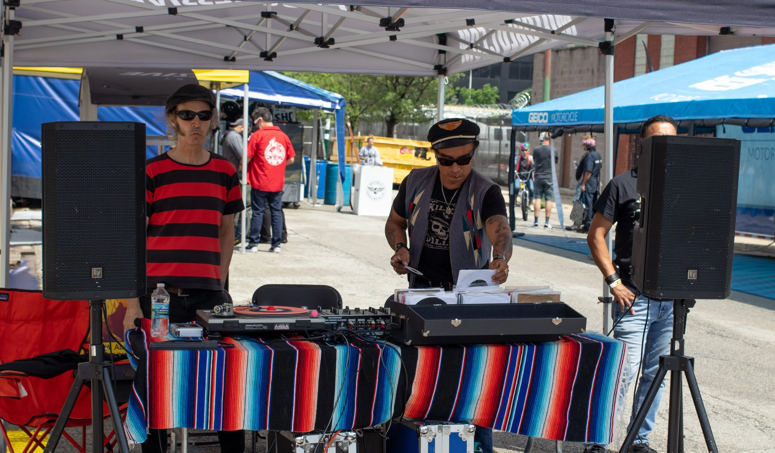 Two men are behind a table with DJ equipment, under a canopy at an outdoor event. One man wears sunglasses, a black hat, and a black and red striped shirt. The other man wears sunglasses, a black cap, and a black shirt. There are speakers, a water bo