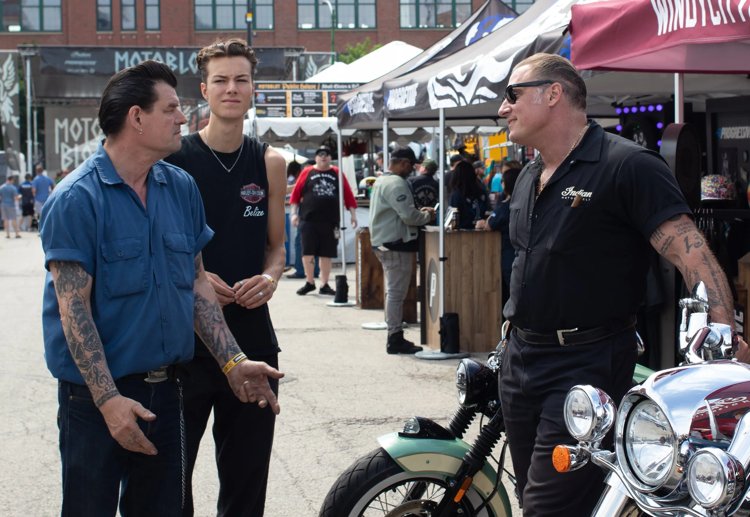 Three men near motorcycles at an outdoor event, with tents and booths in the background.