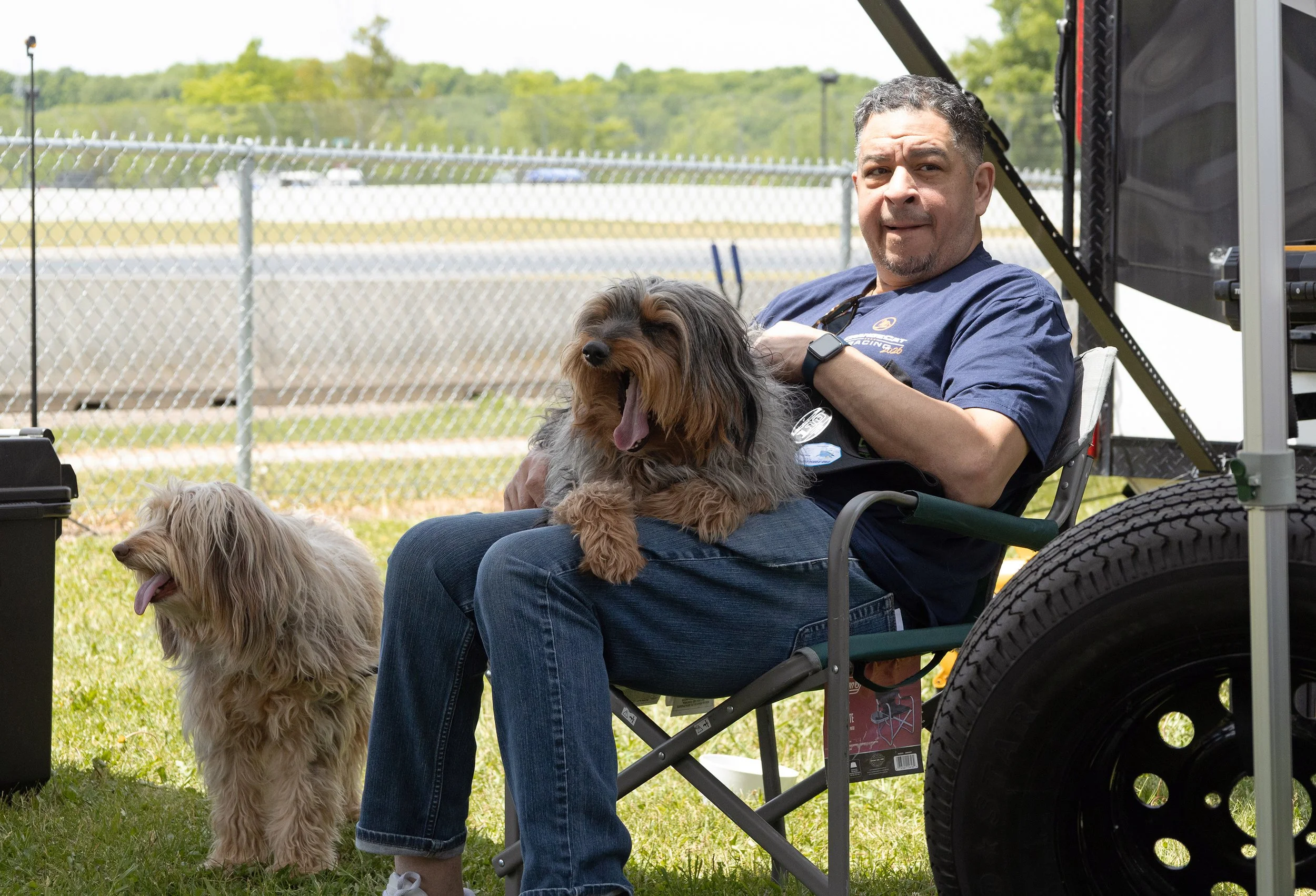 Man sitting in a wheelchair outdoors with two dogs, one on his lap and one standing nearby, in front of a chain-link fence on a sunny day.