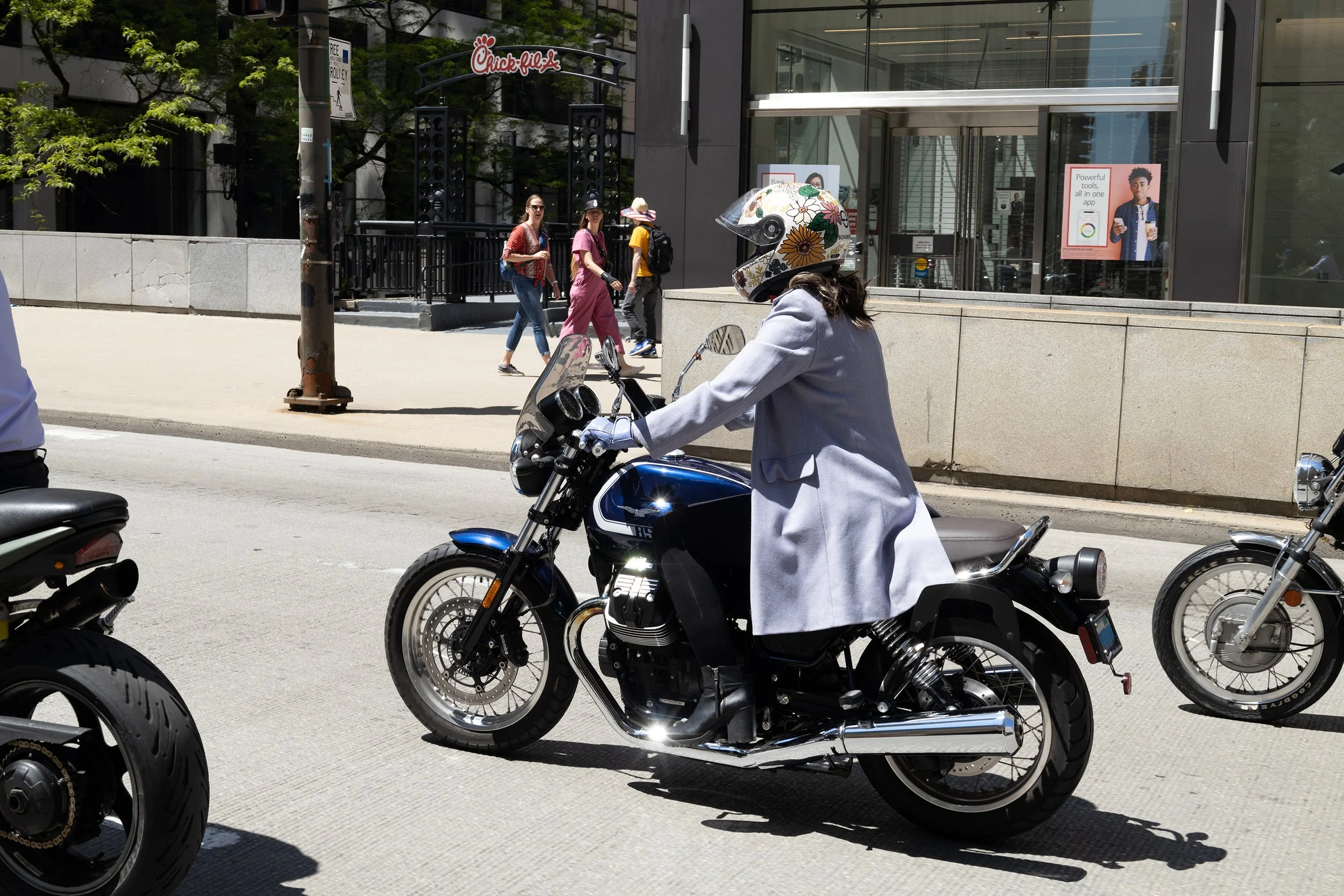 A woman wearing a floral helmet and gray coat riding a black motorcycle on a city street, with pedestrians walking past a Chick-fil-A restaurant in the background.