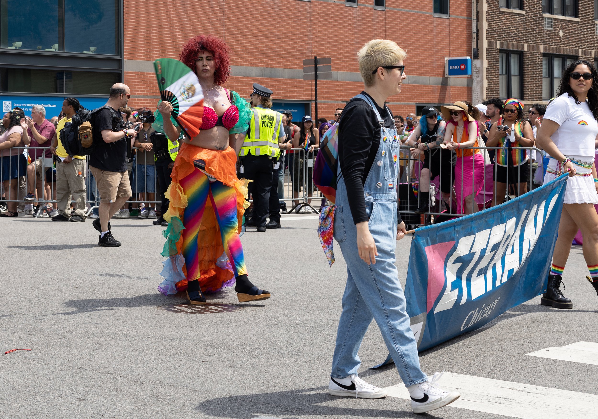A diverse group of people participating in a pride parade. One person is wearing a rainbow-themed outfit with rainbow-colored rainbow leggings and rainbow flags painted on their body, holding a rainbow fan. Others are dressed in casual and colorful o