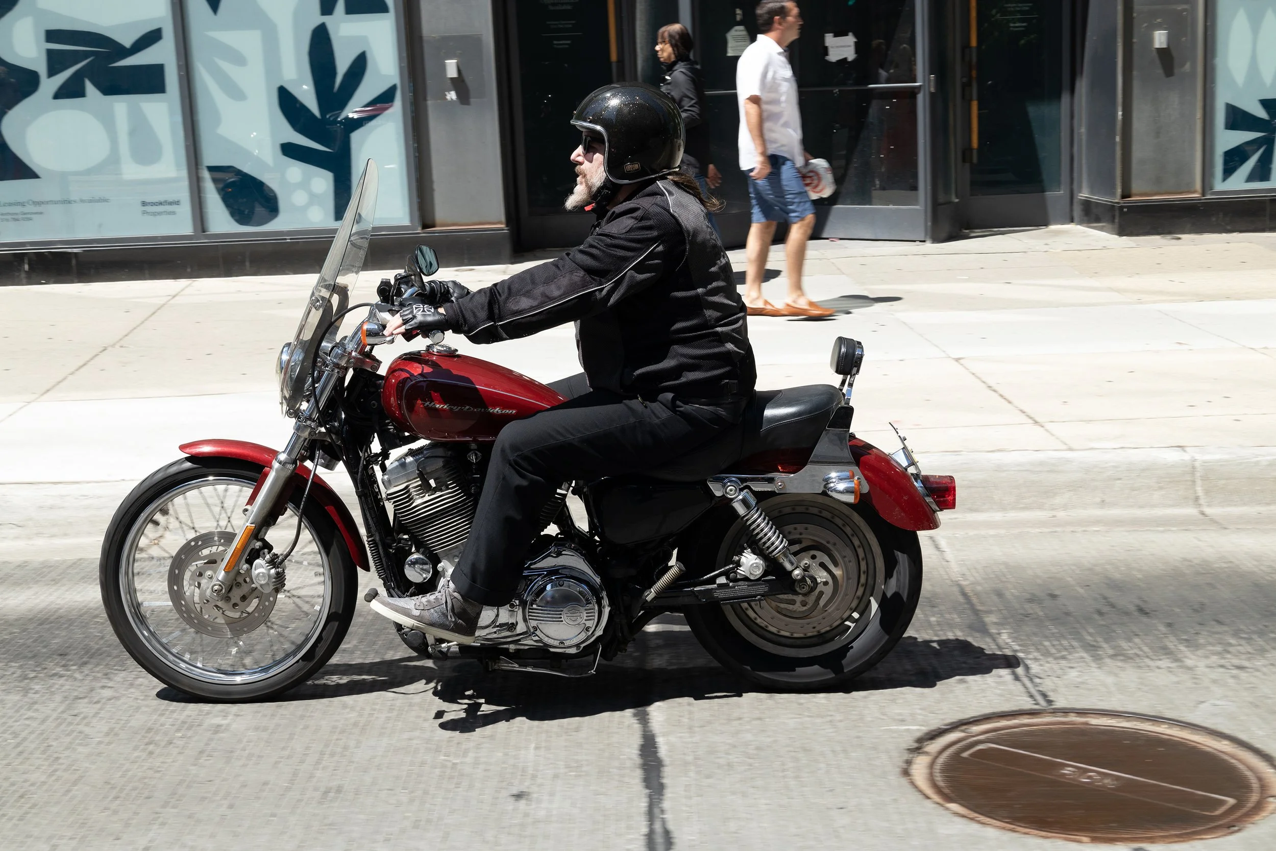 A person with long hair, beard, and glasses riding a red Harley Davidson motorcycle on a city sidewalk, wearing a black helmet, black jacket, and sneakers. Two pedestrians walk past storefronts in the background.