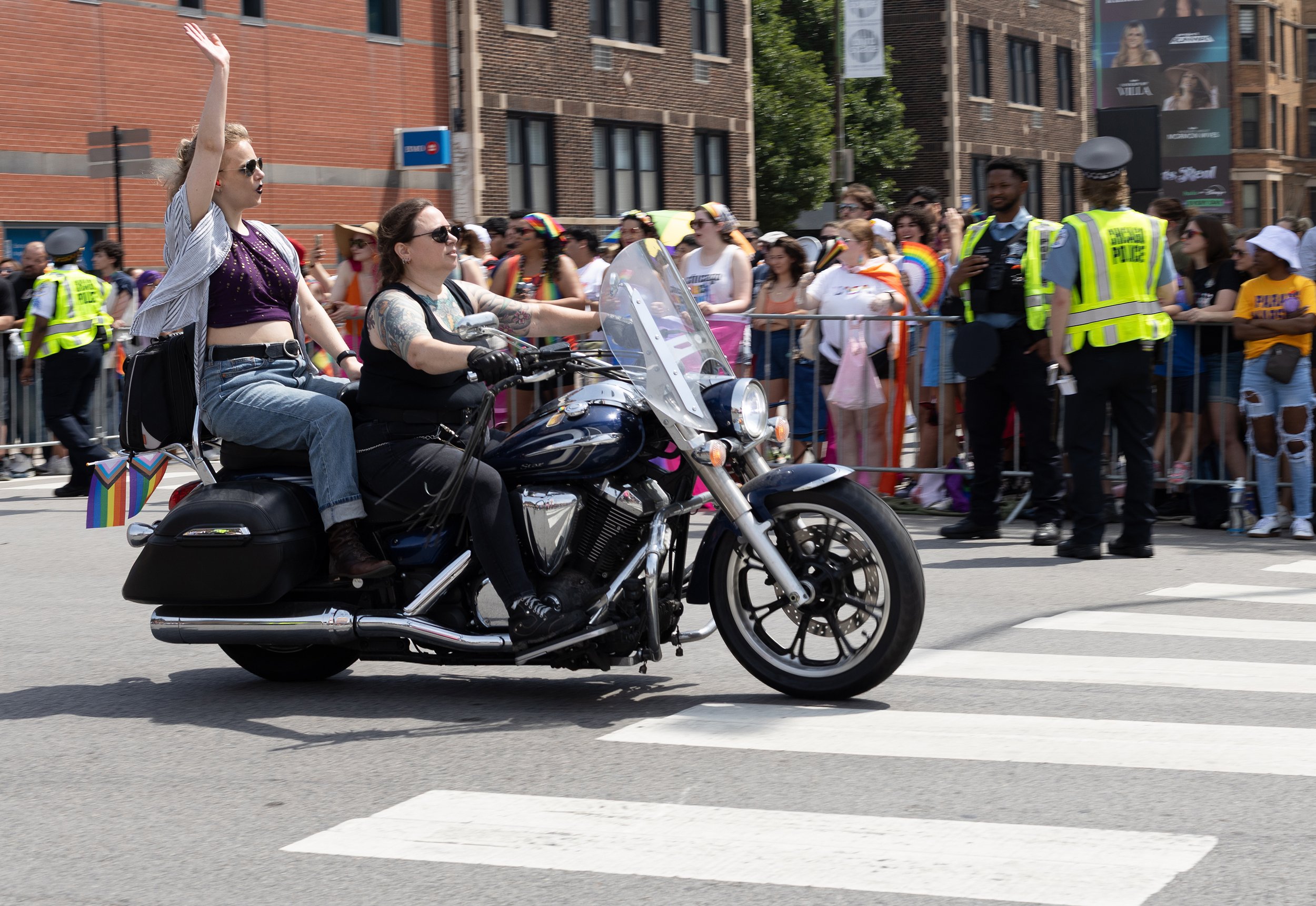 A person riding a motorcycle on a street during a parade, with a crowd of spectators behind a barrier, some wearing rainbow-colored accessories, and police officers present.