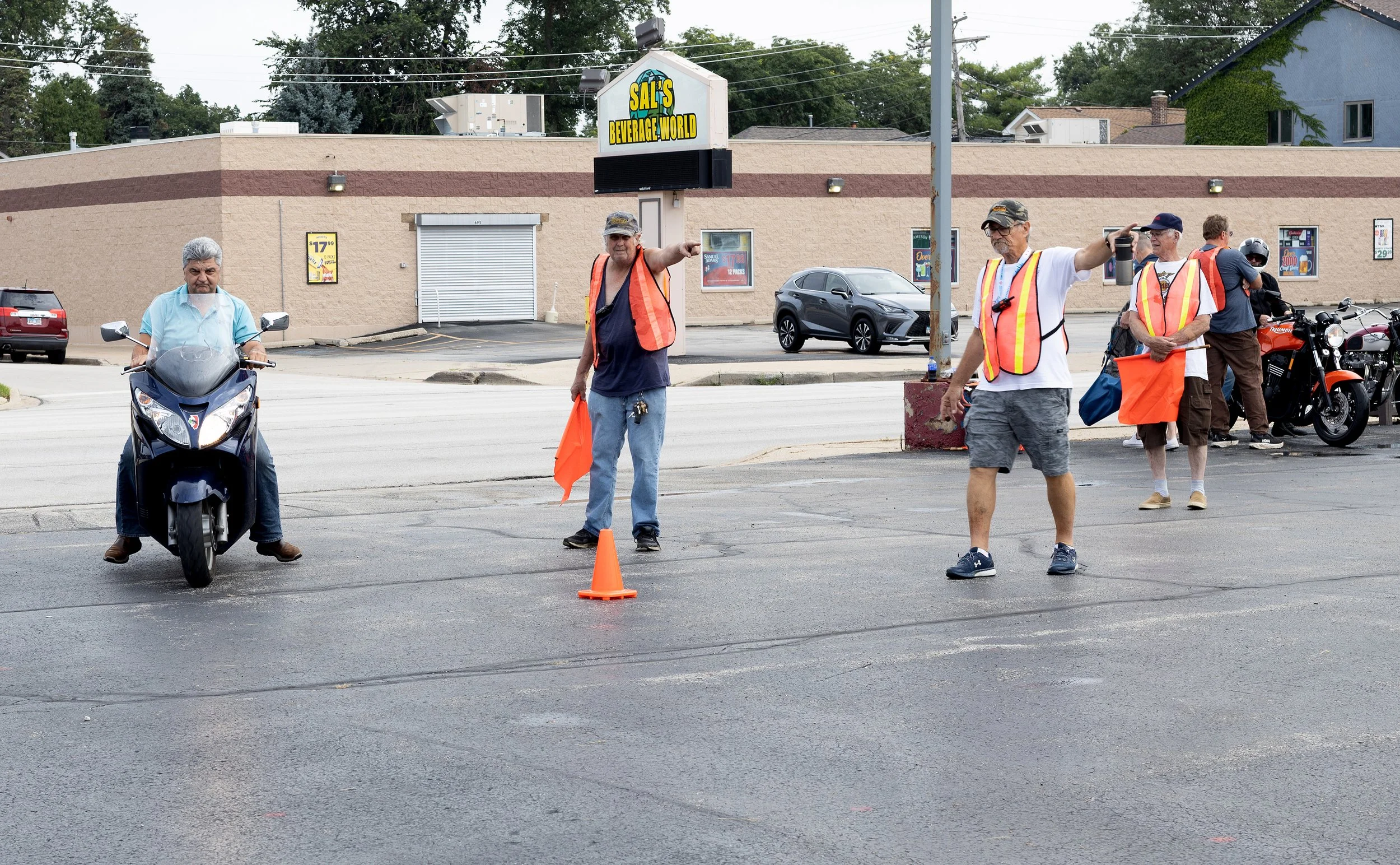 A group of people standing on a street, some with orange safety vests, near a traffic cone, with a building and cars in the background.