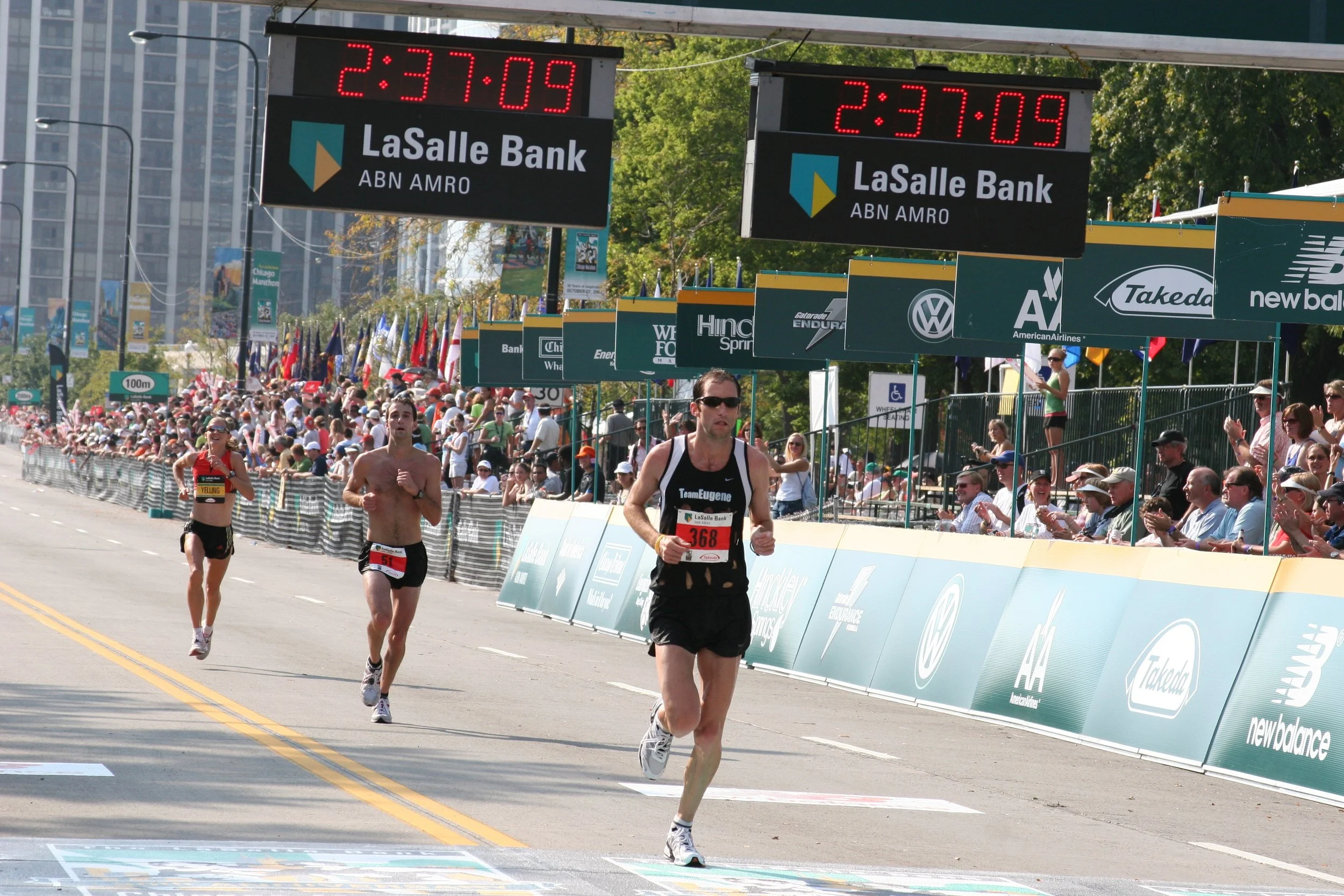 A group of marathon runners competing in a race on a city street, with spectators watching behind barricades, and digital race timers displaying 2:37:09.