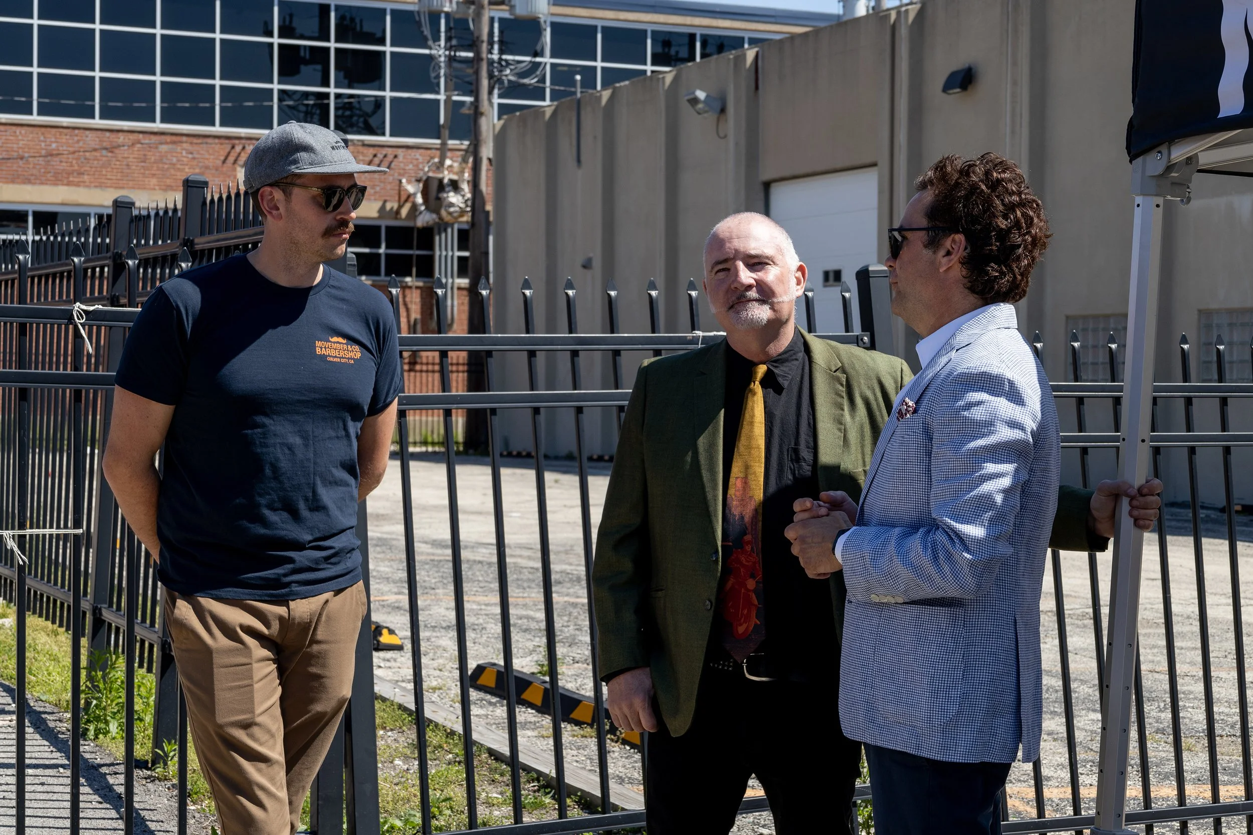 Three men are standing outside near a metal fence, engaged in conversation. One is wearing a navy t-shirt, khaki pants, sunglasses, and a gray cap. The second man is dressed in a green blazer and black shirt. The third is in a light blue checked blaz