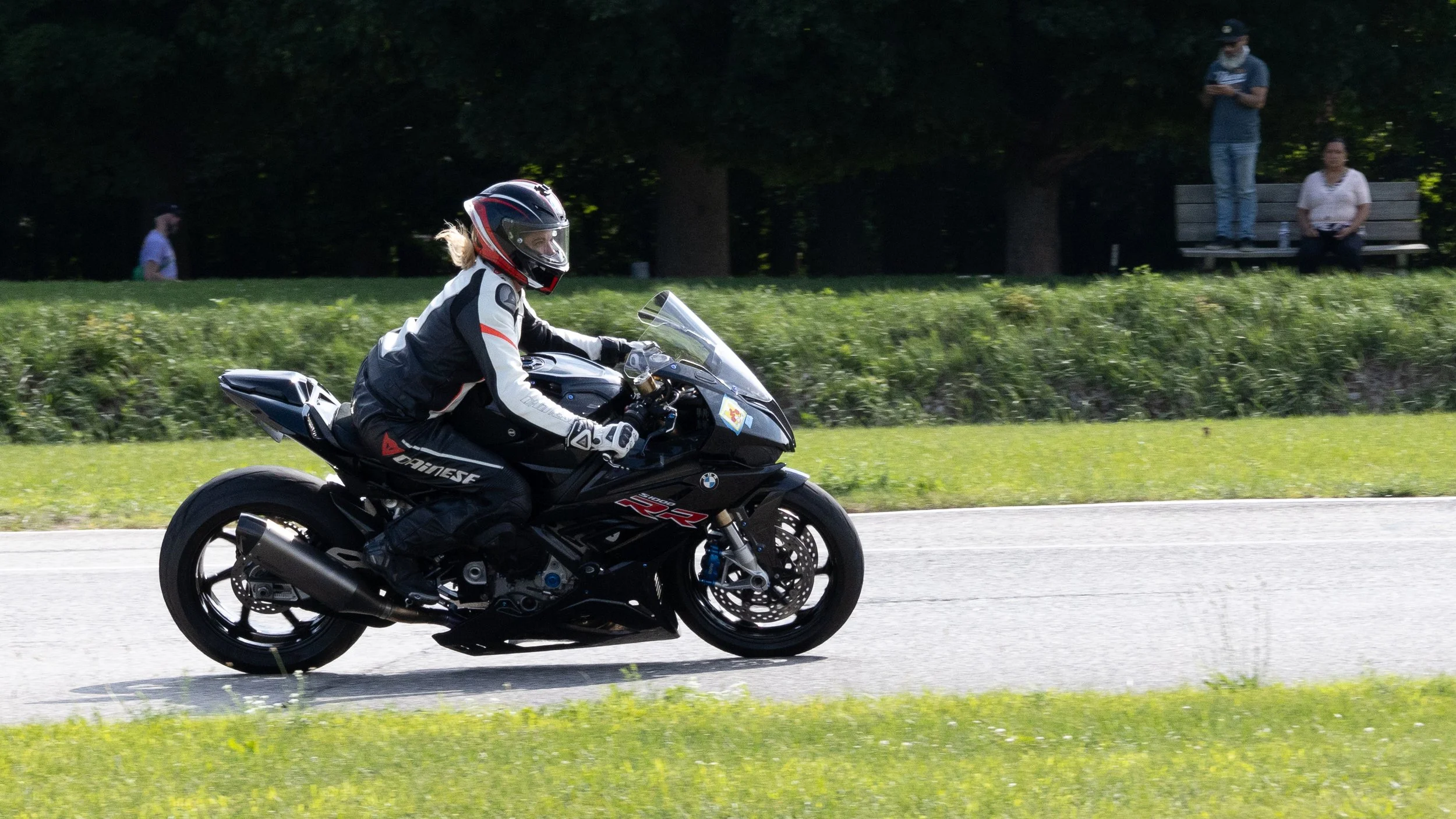 A woman wearing a motorcycle helmet and riding gear is riding a black BMW motorcycle on a paved road, with grassy area and trees in the background.