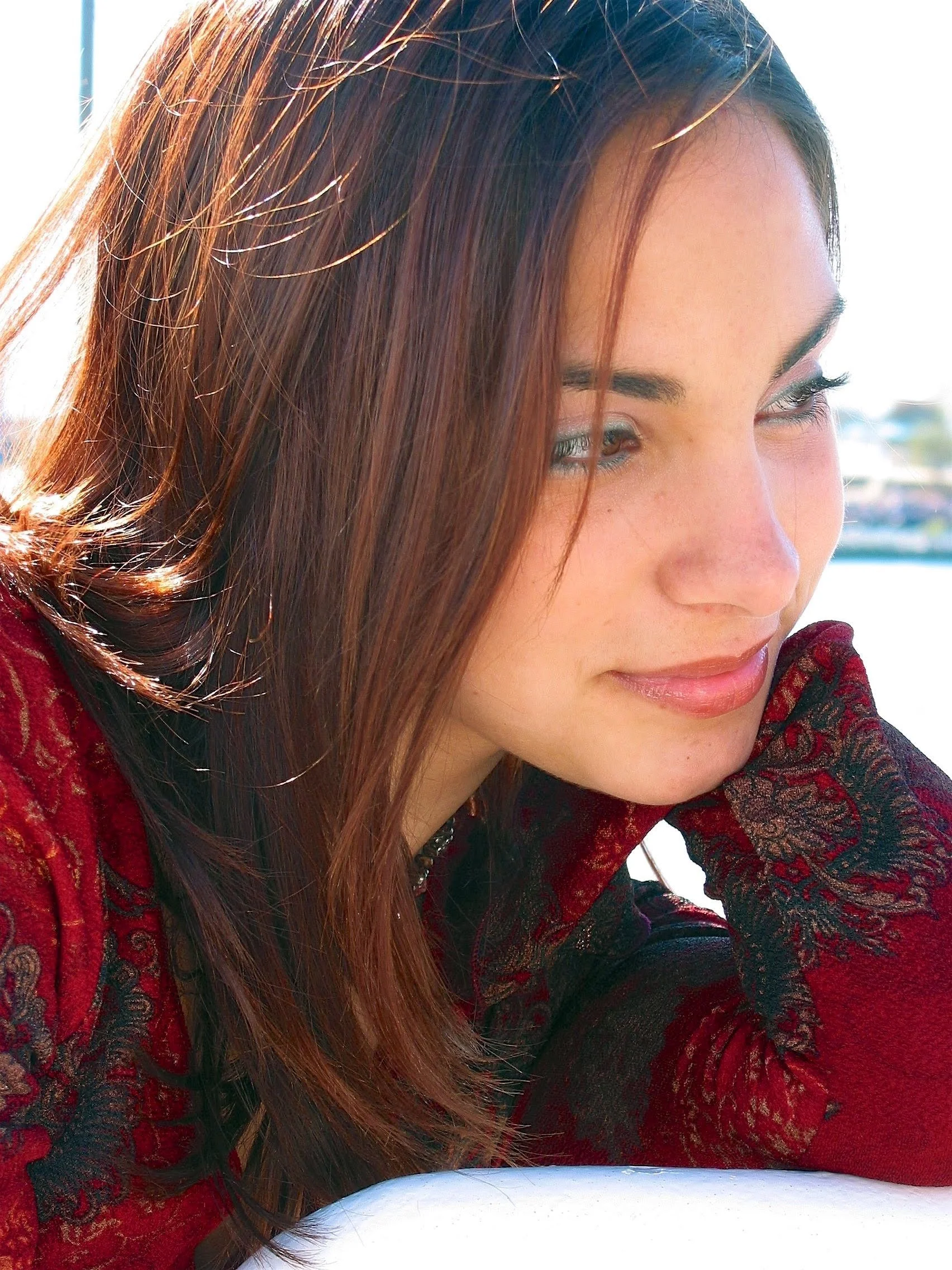 A young woman with long, reddish-brown hair gazes downward, wearing a red and black patterned top, outdoors with bright sunlight and blurred background.