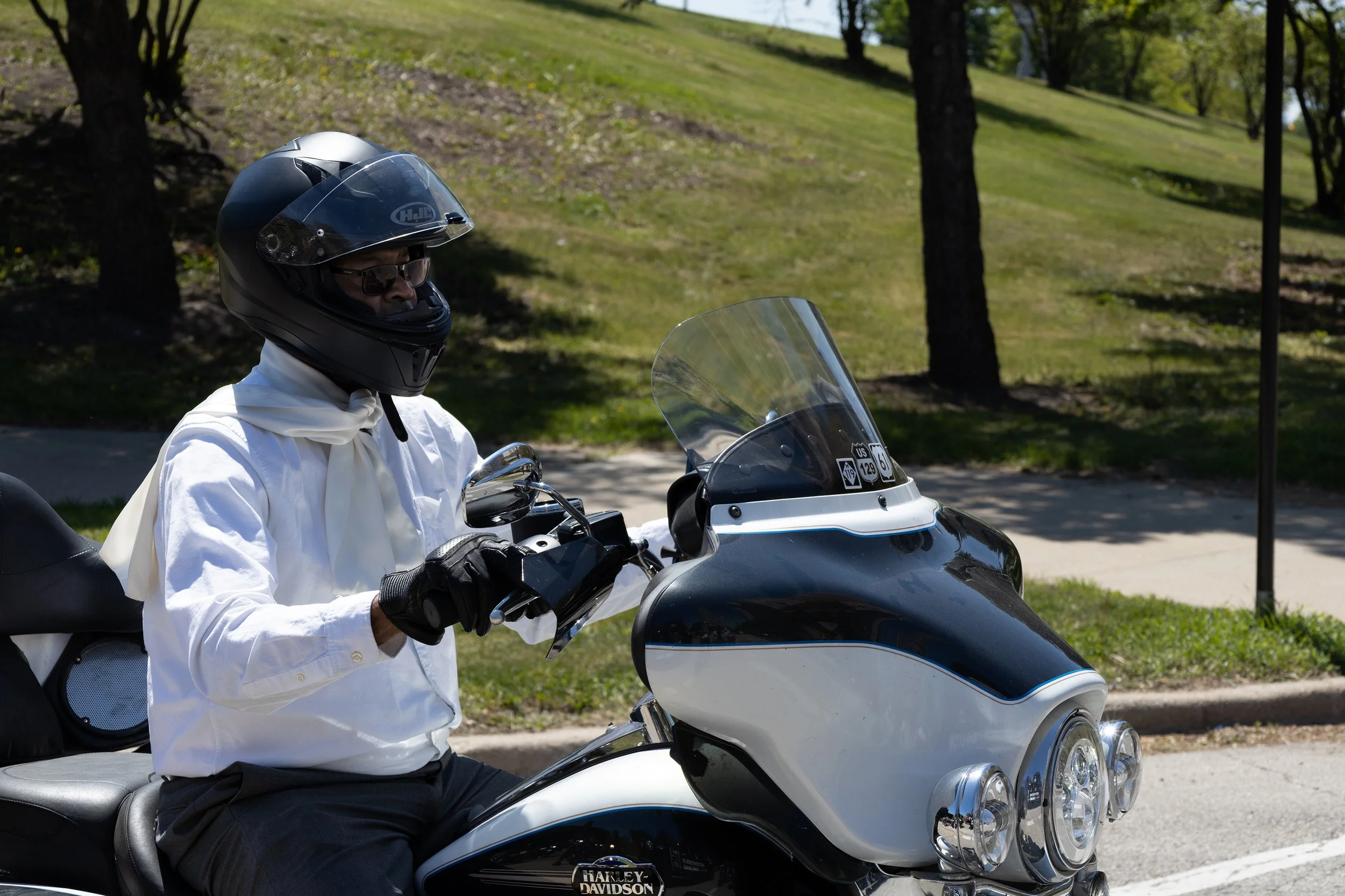 A person wearing sunglasses, a black helmet, a white shirt, and black gloves sitting on a black and white Harley-Davidson motorcycle on a street with green trees and grassy hill in the background.