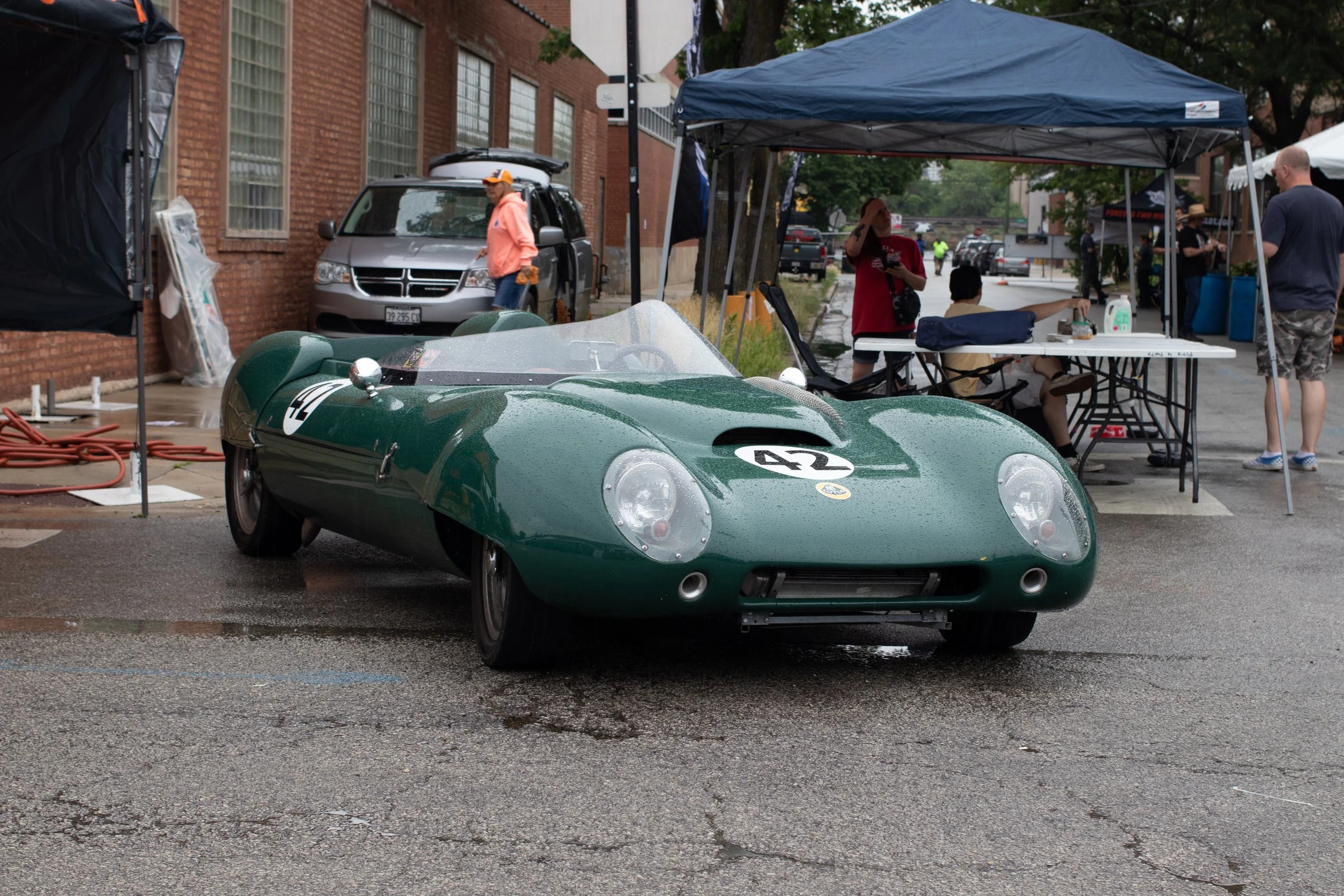 A vintage green race car with the number 42 on its hood parked on a wet street during a car event, with tents and people in the background.