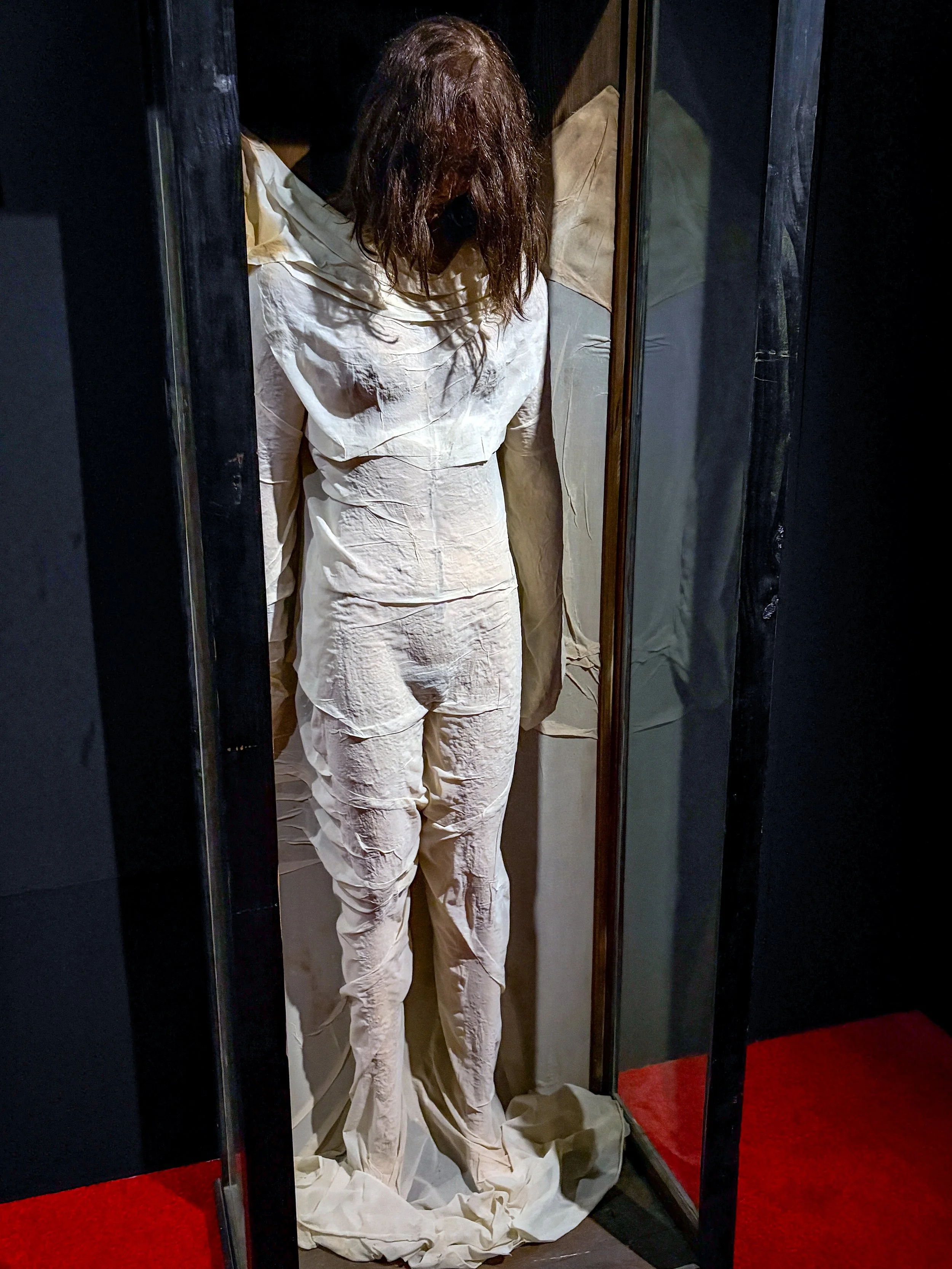 A person with long, wavy hair wearing white pajamas standing inside a small enclosure with black frames and mirrors, with a red carpet on the floor.