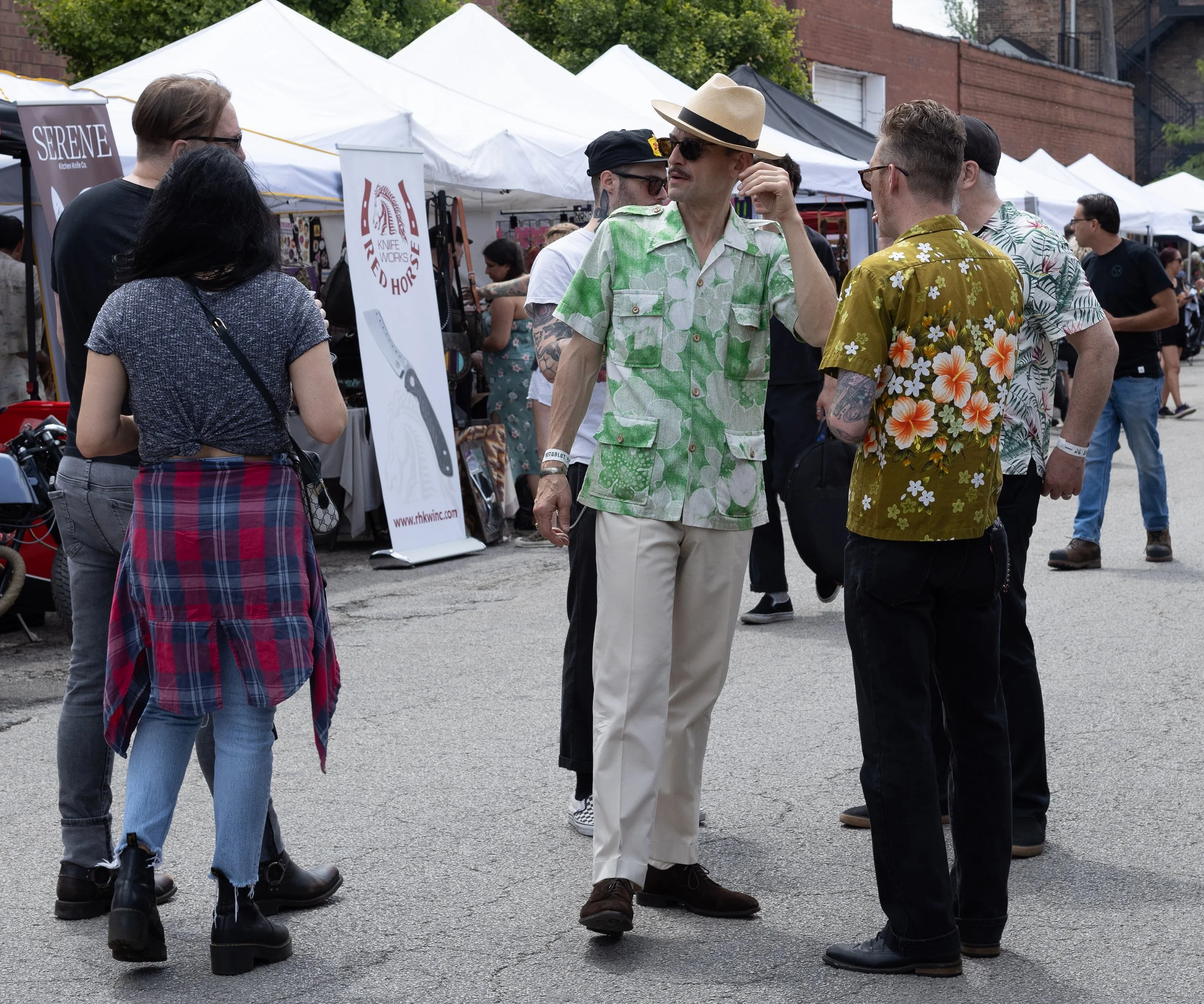 A group of people conversing outdoors at a street market or festival with tents and vendor signs in the background. The group includes a man wearing a straw hat, sunglasses, a green floral shirt, and light-colored pants, talking to two men wearing fl
