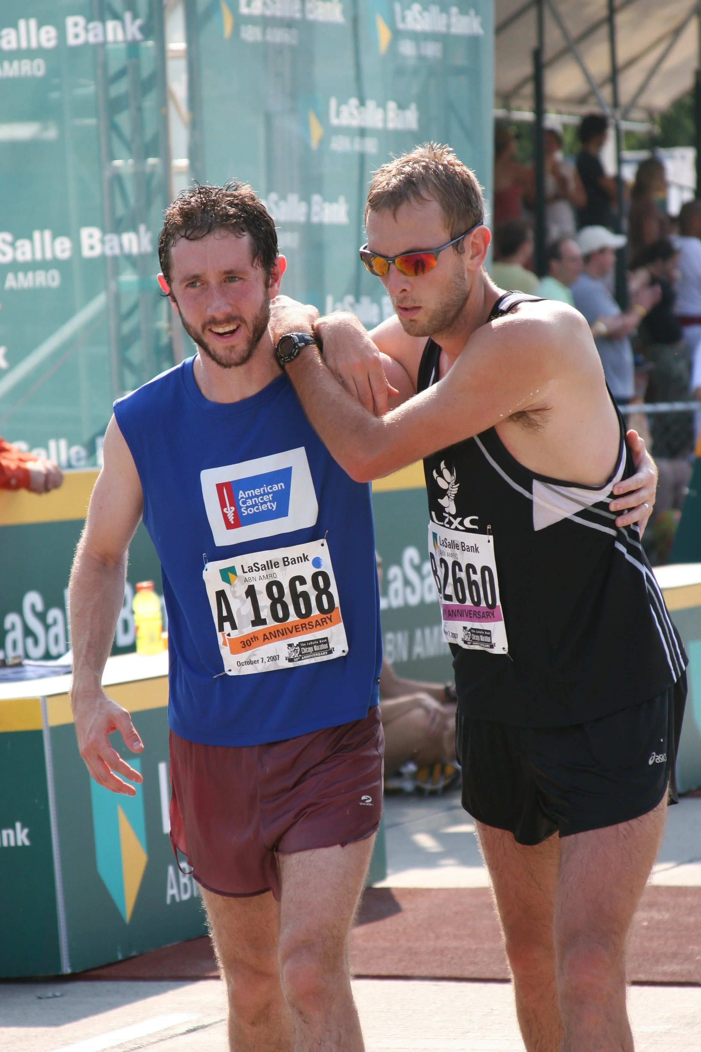 Two male runners are seen after completing a race, with one helping the other. Both are wearing athletic clothing, race bibs, and sunglasses. The background shows a crowd and banners at the race event.