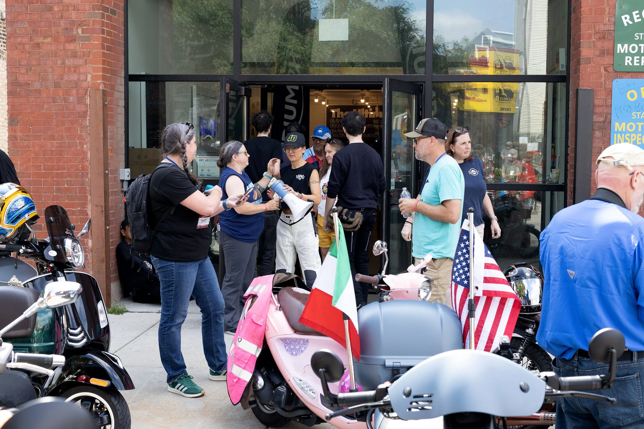 People gathered outside a storefront, some talking and walking among parked scooters decorated with flags, including American and Italian flags. The setting appears lively with motorcycle enthusiasts or a similar event happening.