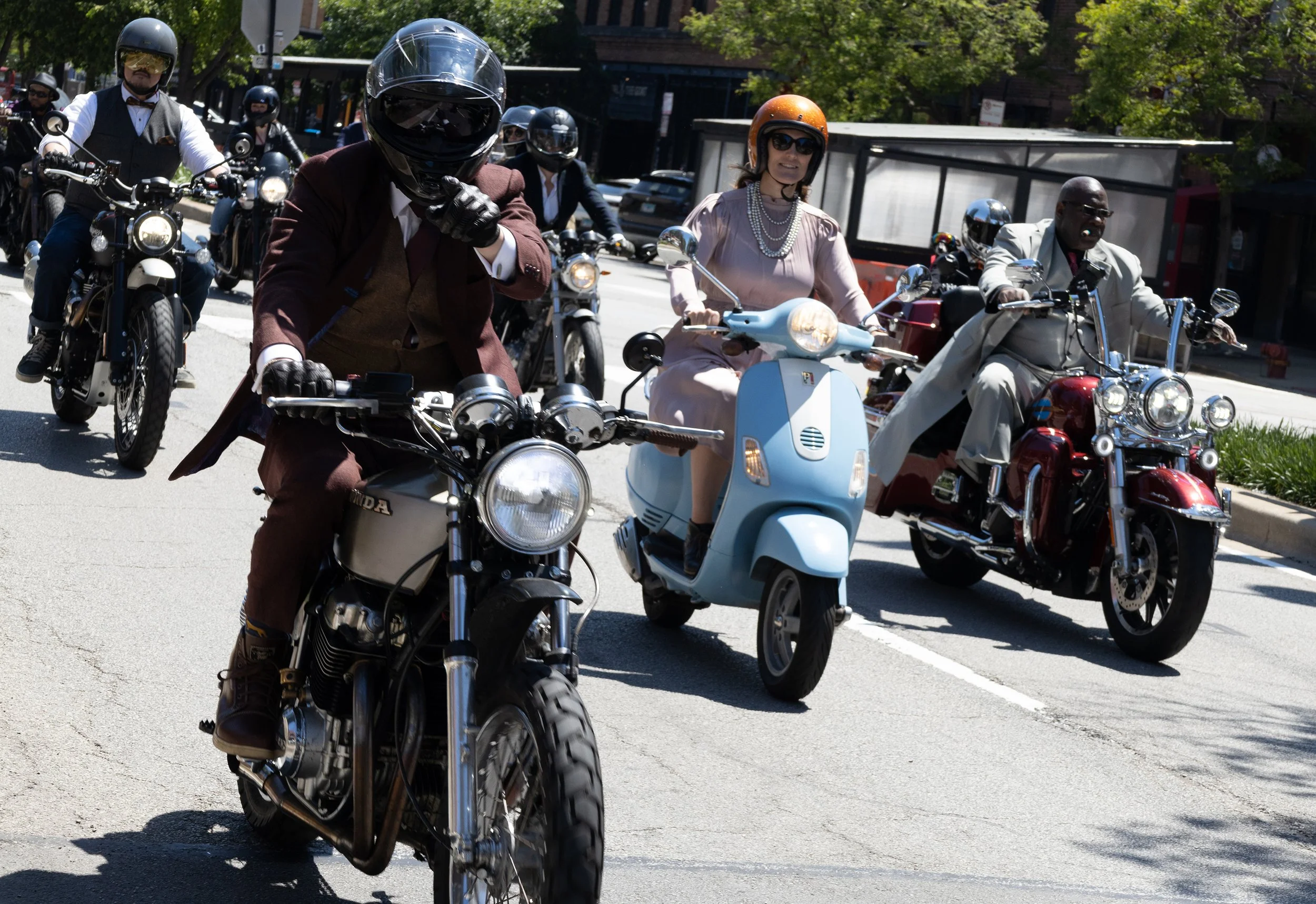 Group of people riding motorcycles and a scooter on a city street during daytime, wearing helmets and vintage or formal clothing.