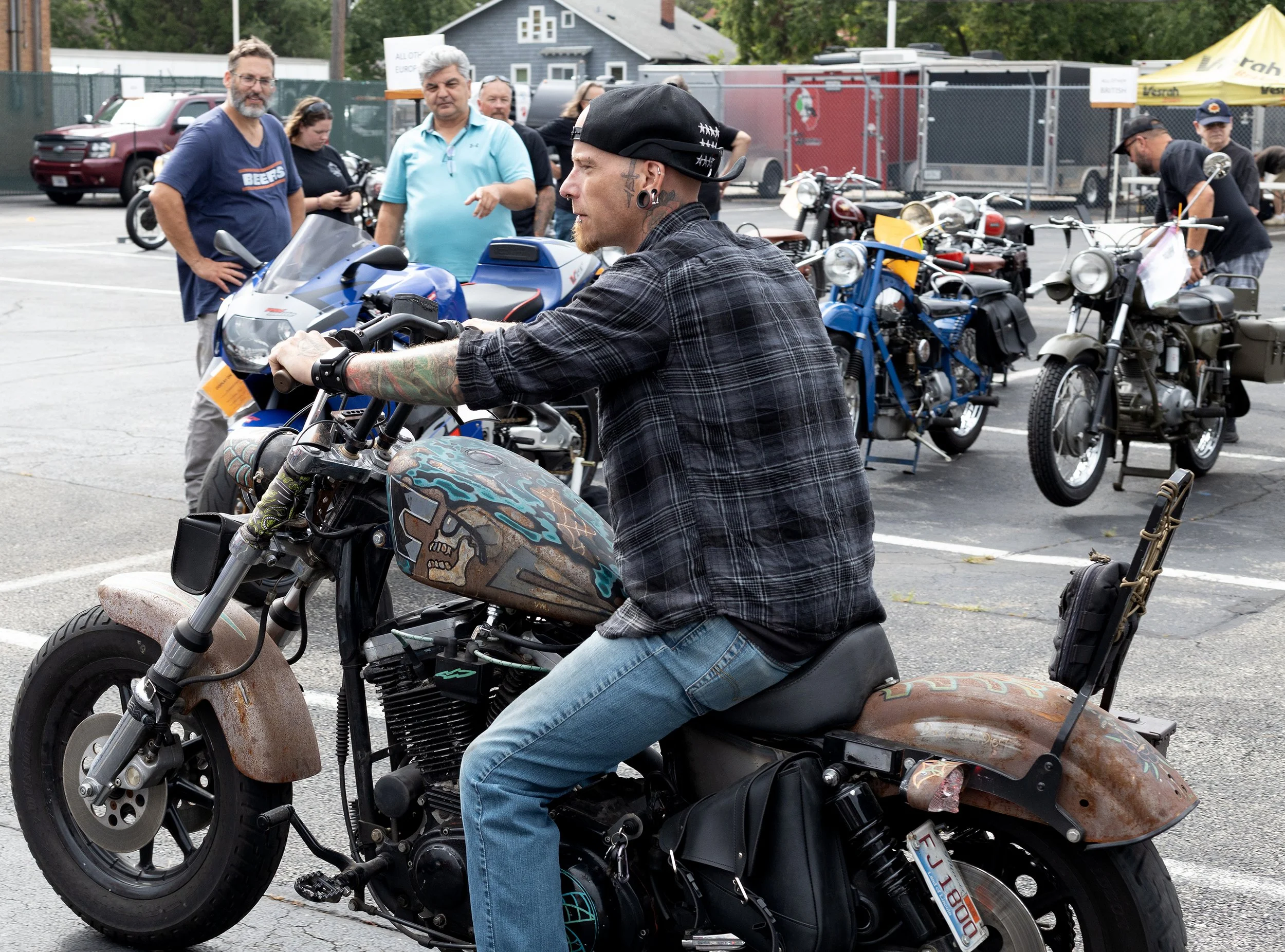 A man with tattoos and a backward cap is sitting on a custom motorcycle at a motorcycle gathering, with people and bikes in the background.