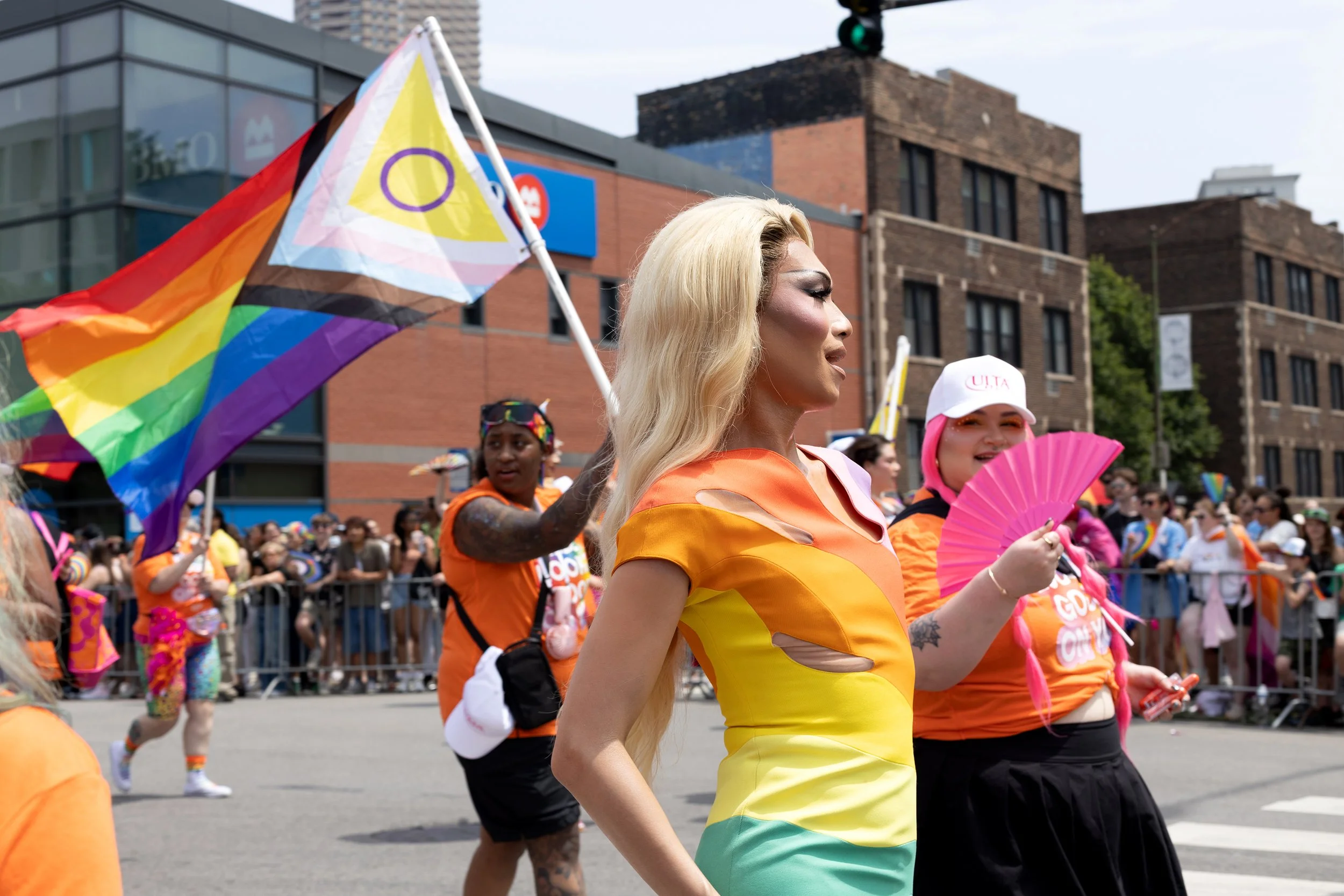 A person with long blonde hair and makeup, wearing a rainbow-colored dress, participating in a parade with others dressed similarly, holding a rainbow flag, with a crowd and city buildings in the background.
