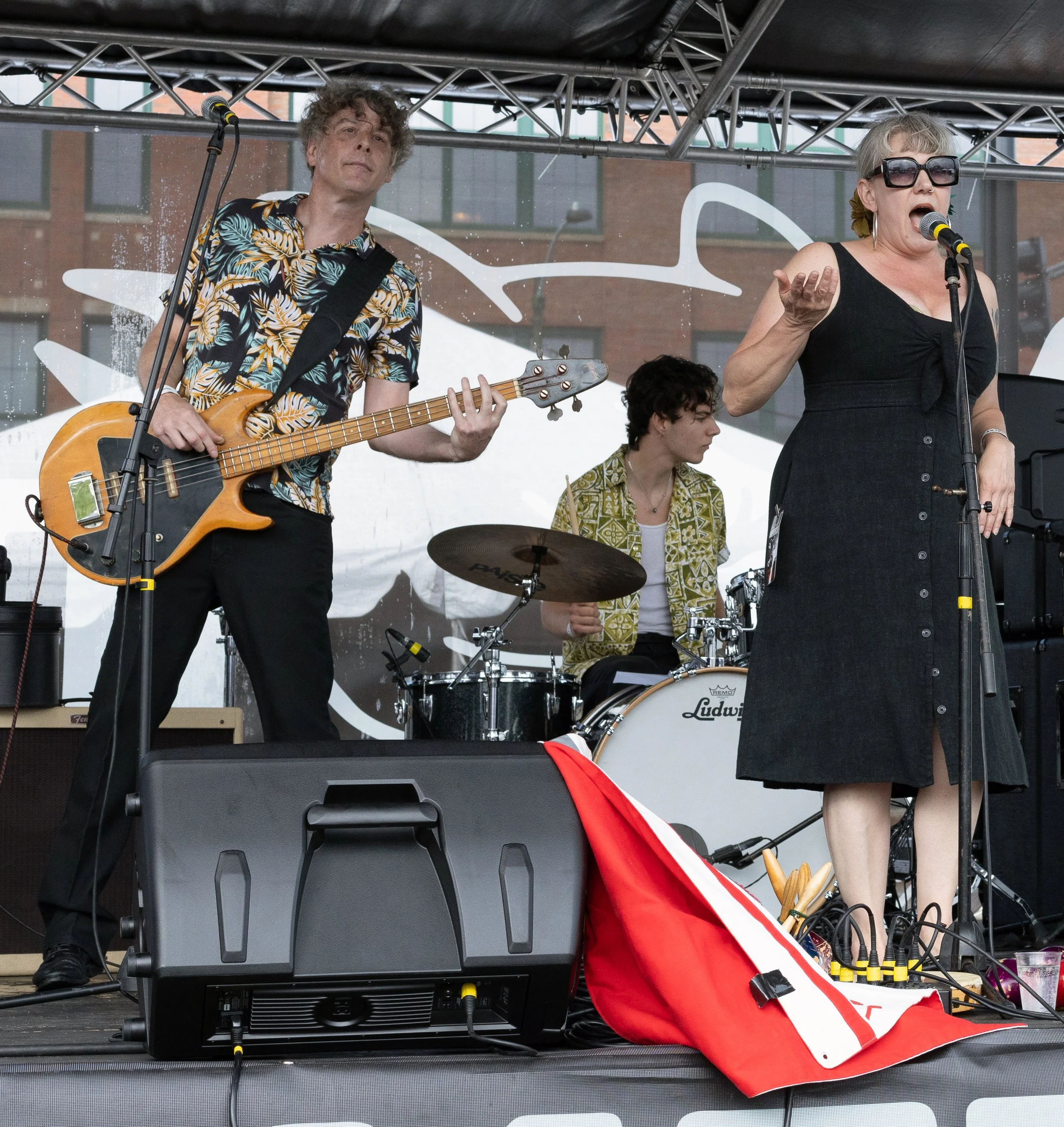 A band performing on an outdoor stage with a guitarist, drummer, and singer, with a large sign in the background. The guitarist is wearing a colorful, floral shirt and playing an electric bass guitar, while the singer is dressed in a black dress, lar