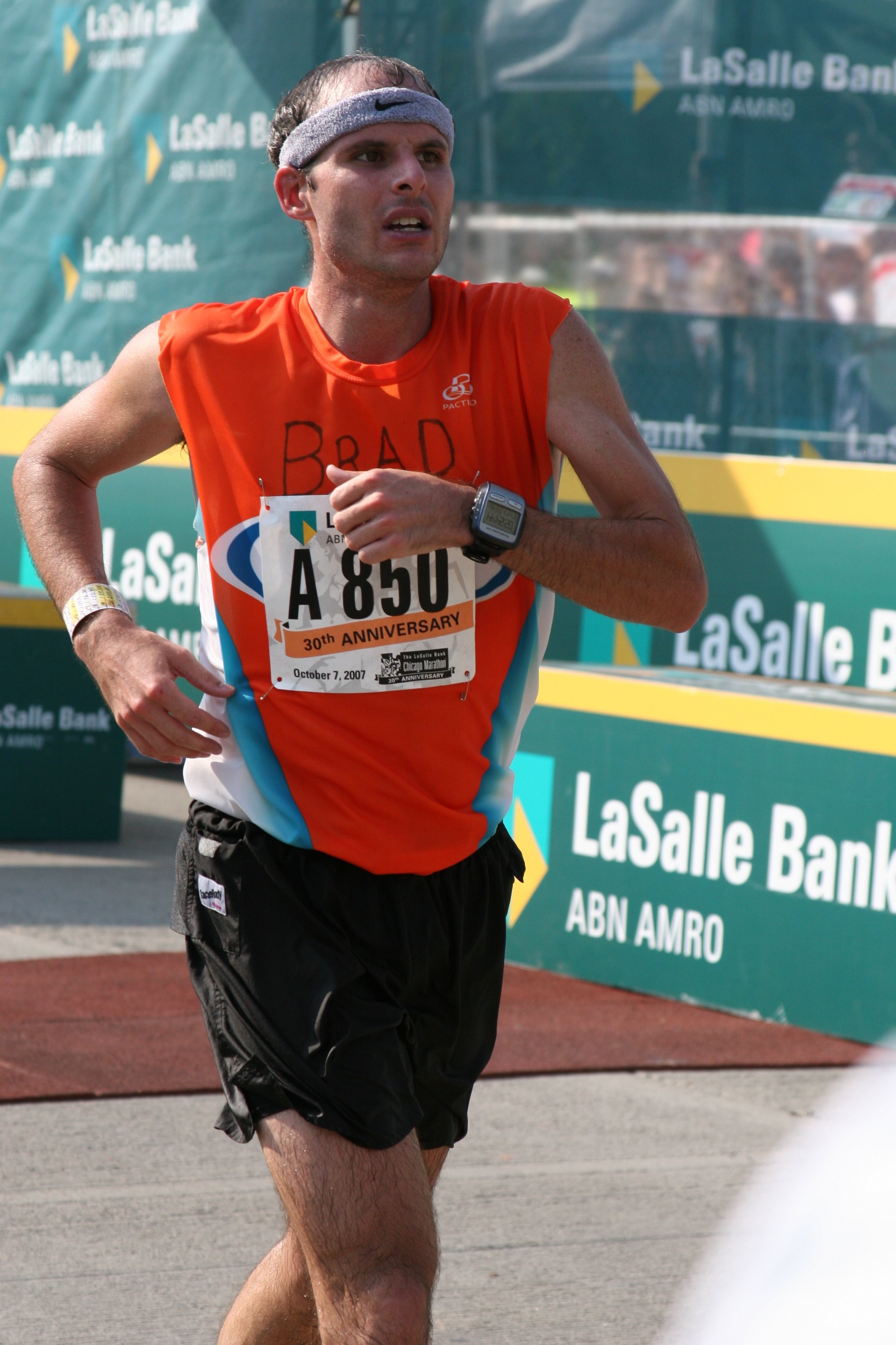 A male runner wearing an orange and blue vest, black shorts, and a gray headband running in a marathon with a race bib that reads 'A 850' and '30th ANNIVERSARY'.
