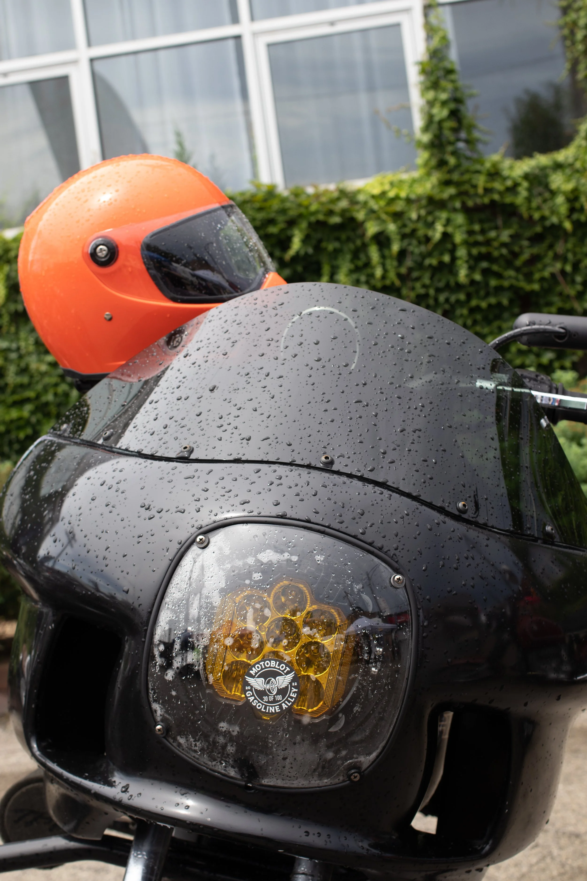 Black motorcycle with a black headlight that has a Motoblot Gasoline Alley sticker on it. An orange helmet is resting on the motorcycle with droplets of water on the helmet and motorcycle. There are green bushes and a glass building in the background