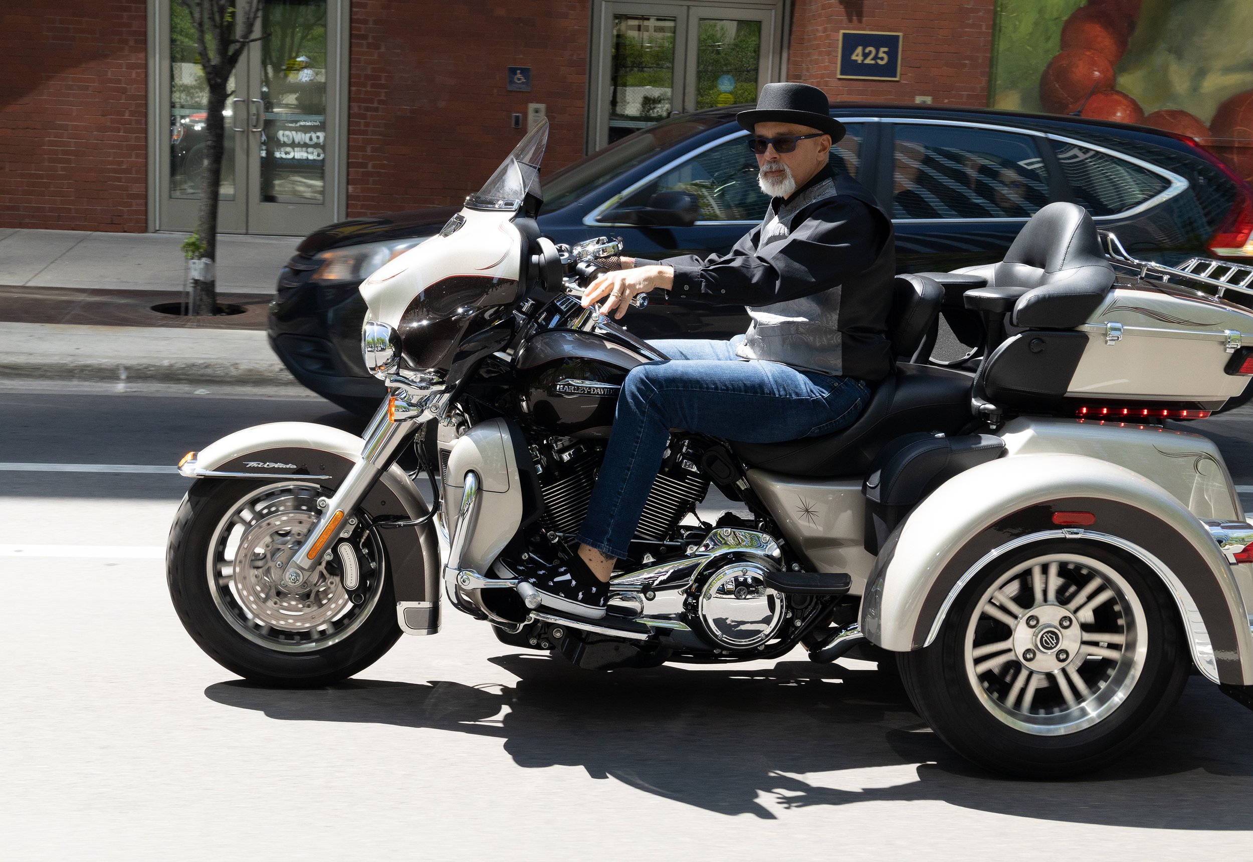 A man wearing sunglasses, a black hat, a black and gray jacket, and jeans is riding a white and black Harley-Davidson motorcycle on the street.