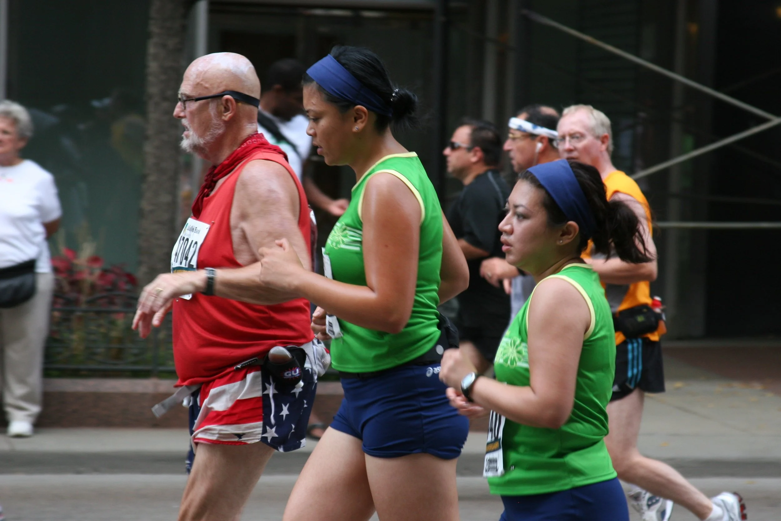 A group of runners participating in a marathon, with two women in green shirts and a man in a red shirt, running on a street with others in the background.