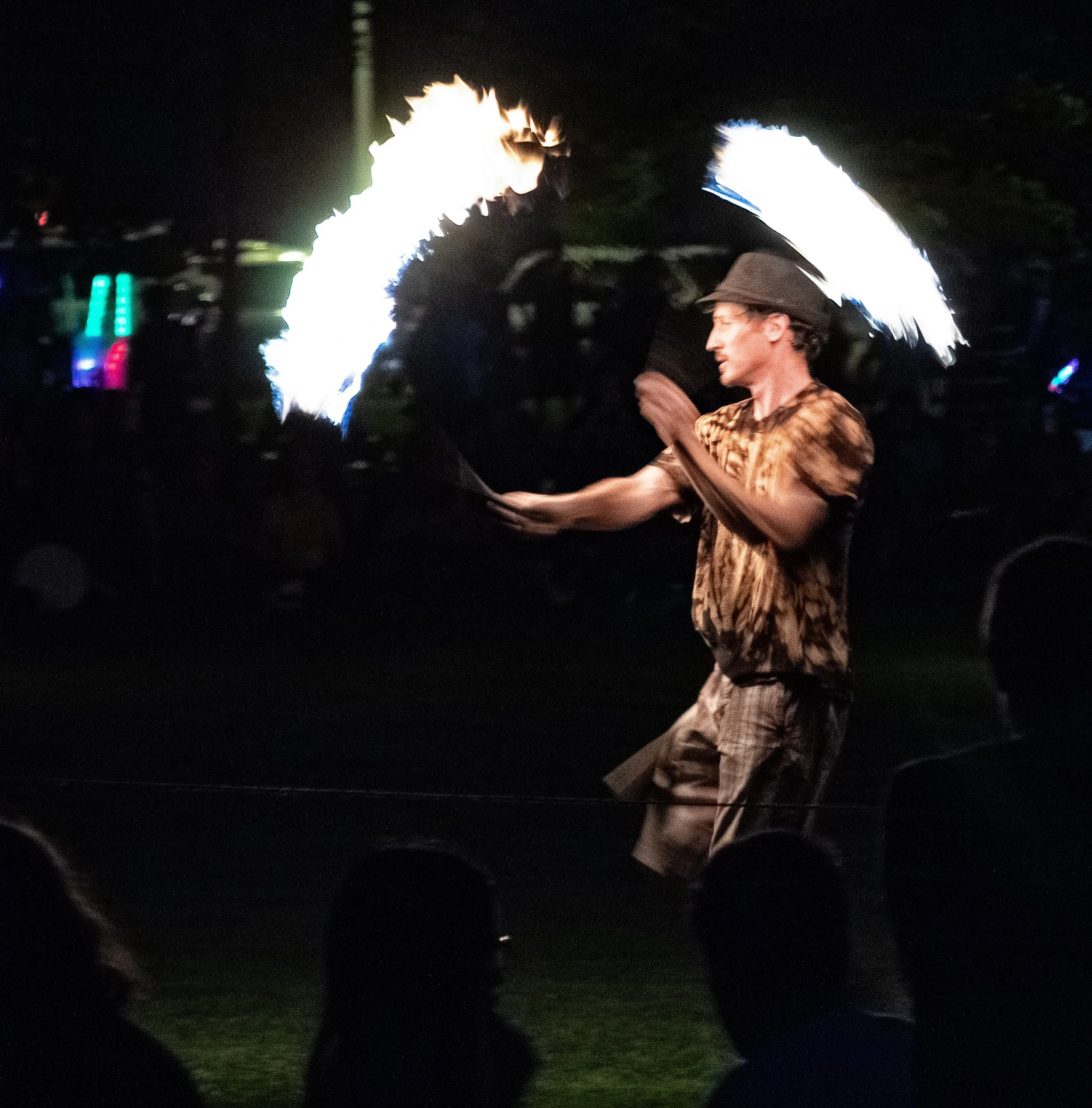 A person with light skin is performing a fire breathing act at night, creating a large flame projected outward. The performer is wearing a dark hat and a patterned shirt, and is smiling with eyes closed. The background is dark with some faint colorfu