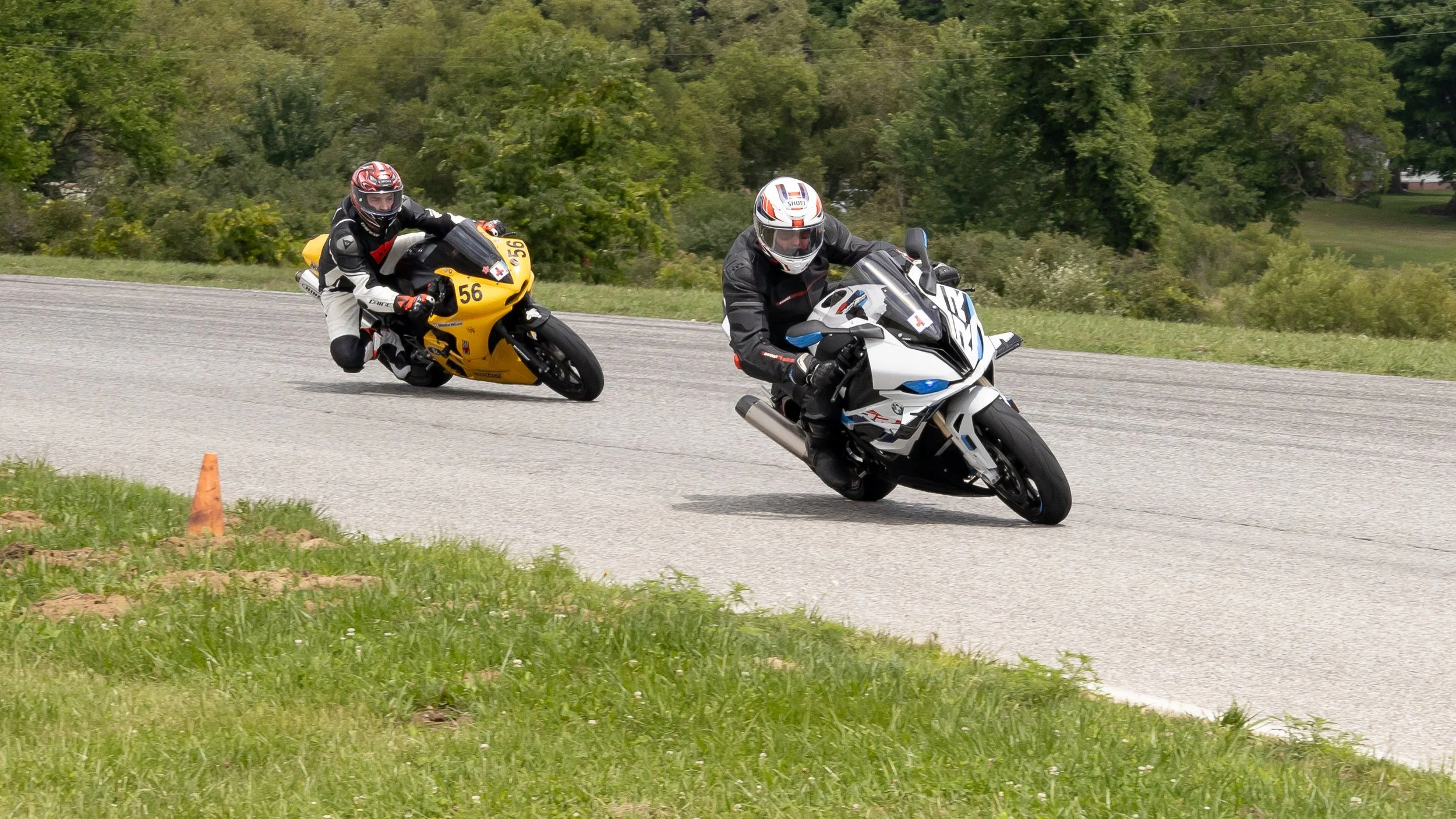 Two motorcyclists racing on a curved road, surrounded by green trees and grass.