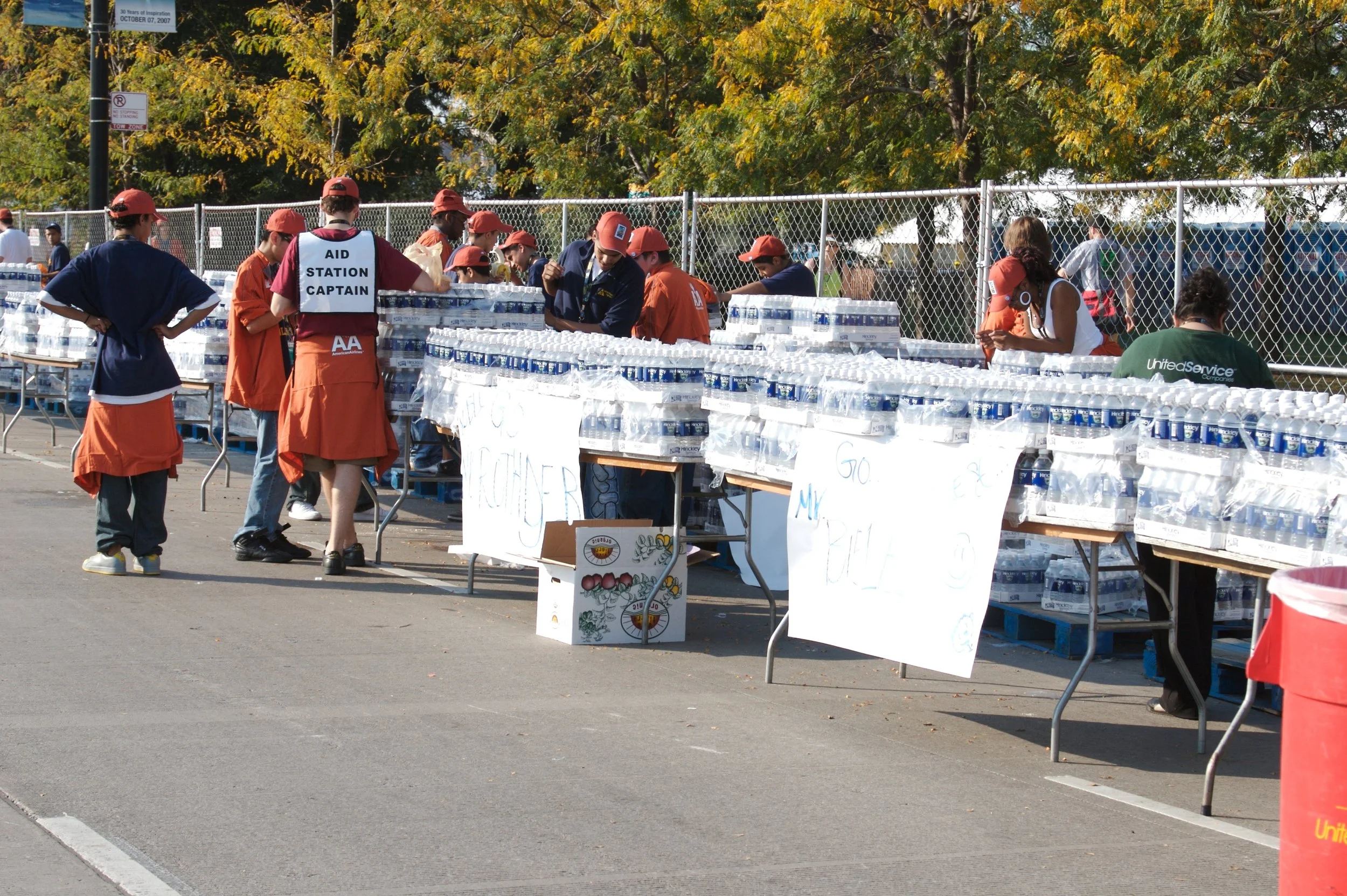 People working at a water distribution station with bottled water, some wearing uniforms and hats, outdoors with trees in the background.