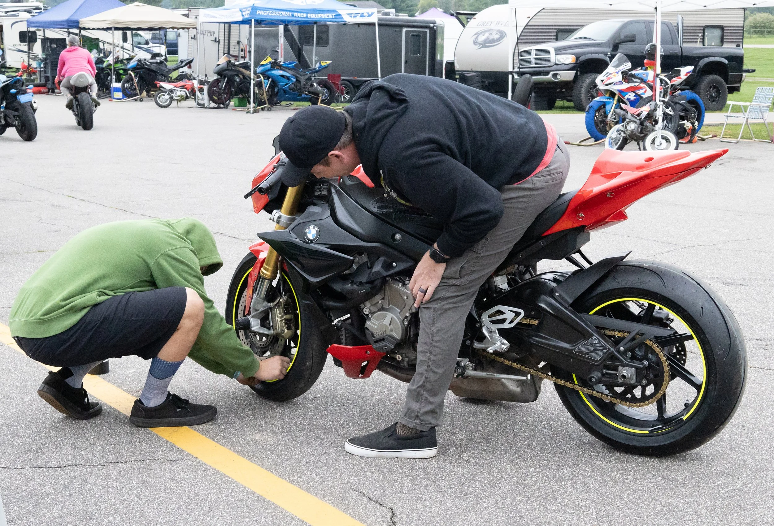 Two men are working on a black and red BMW motorcycle in a parking lot. One man is kneeling, and the other is bending over the motorcycle. The parking lot has other motorcycles, cars, and trailers in the background.