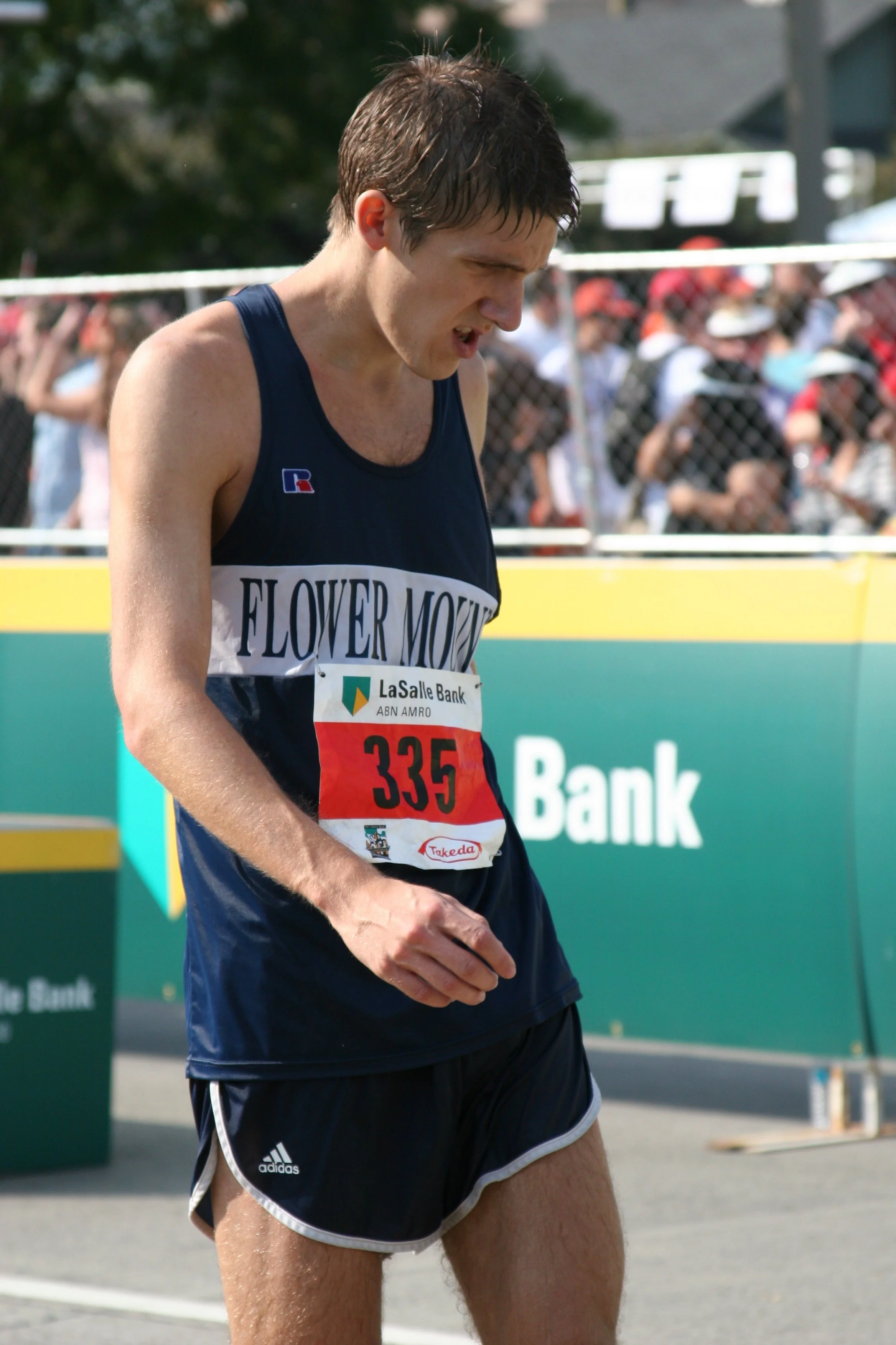 A male track athlete in a dark blue uniform with the text 'FLOWER MOY' on the front, standing near a track with a crowd in the background, appears to be catching his breath after running.