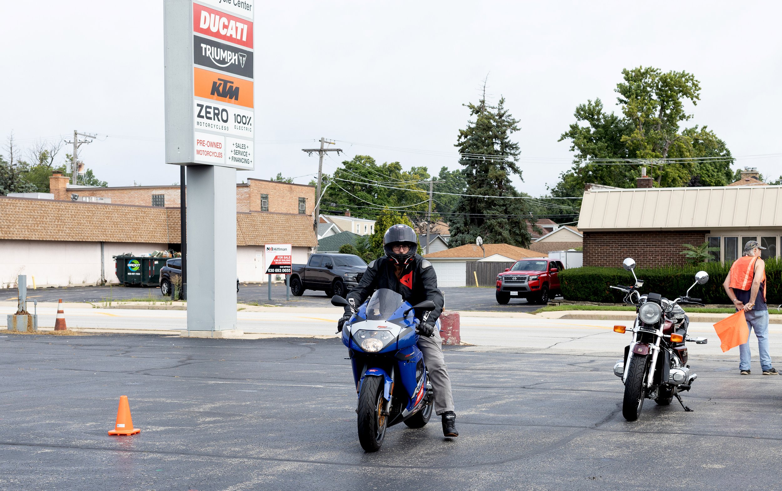A motorcyclist in a black helmet and jacket riding a blue race motorcycle on a paved lot with orange traffic cones. In the background, a person in an orange vest is holding an orange flag, and other motorcycles and cars are parked nearby. There is a 