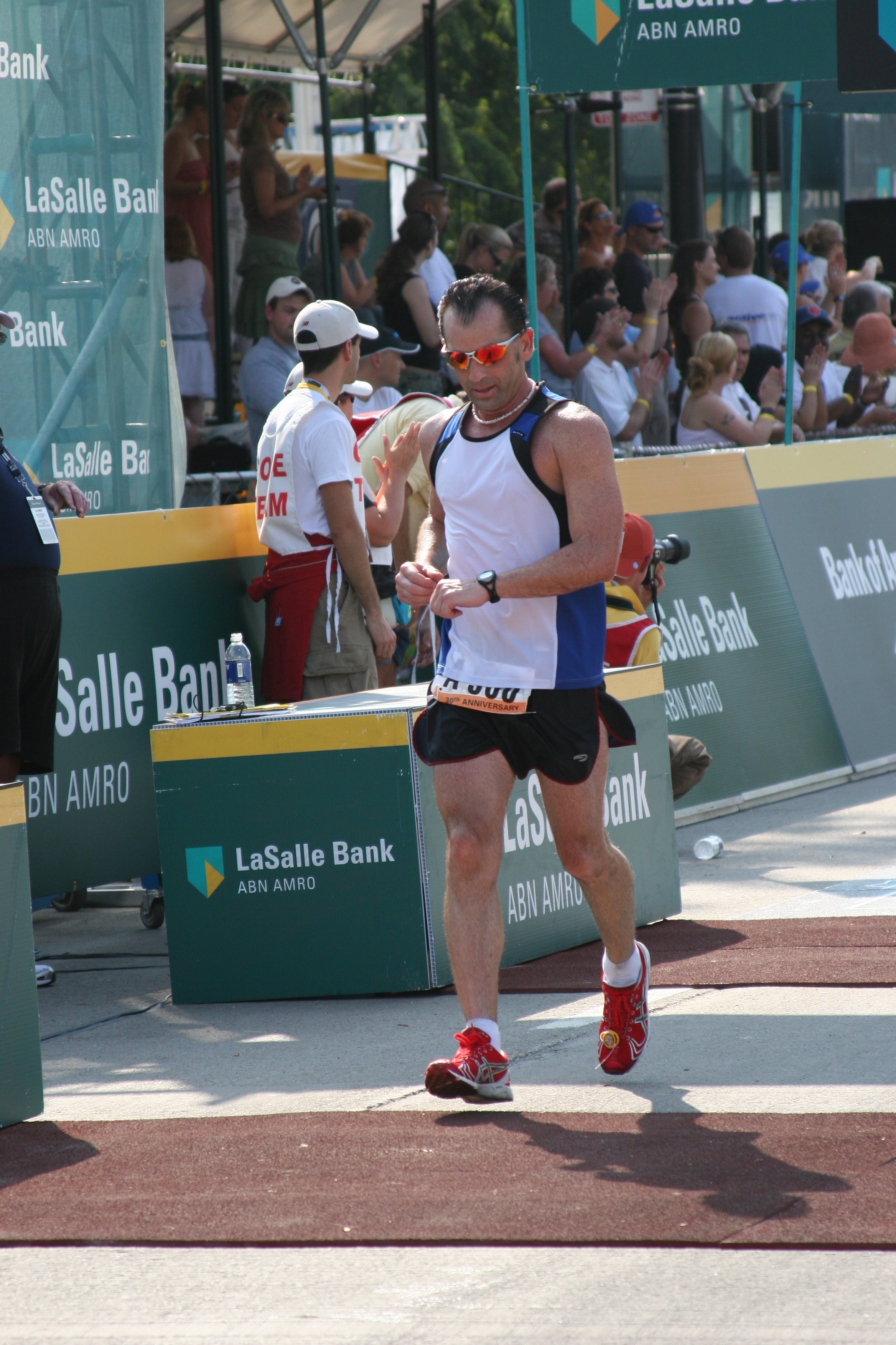 A male marathon runner wearing sunglasses, a white tank top, black shorts, and red running shoes, crossing the finish line with a crowd of spectators clapping in the background.