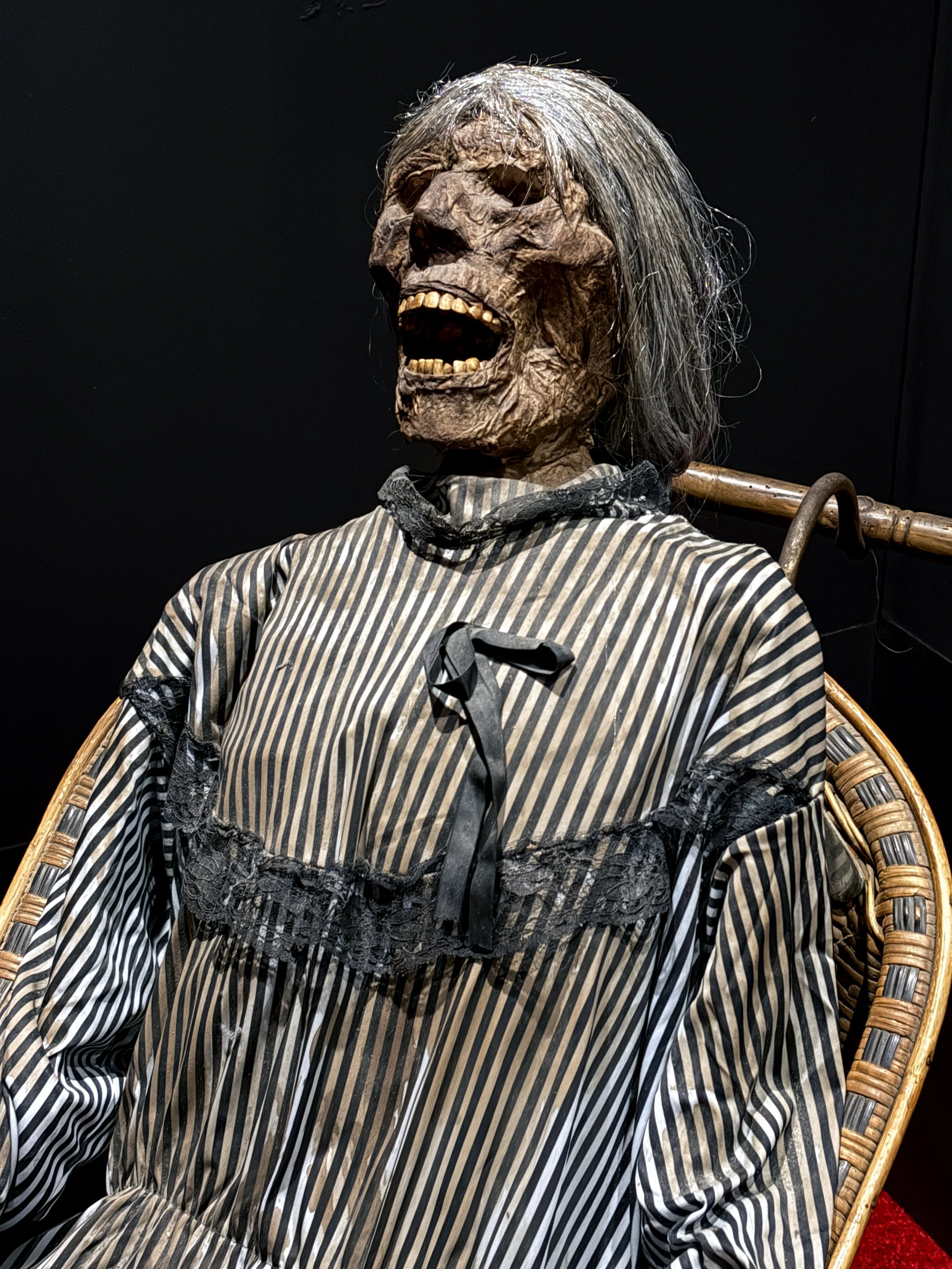 A life-sized humanoid figure with a skeletal, decayed face, hollow eyes, and long gray hair, dressed in a black and white striped dress with lace accents, sitting on a wicker chair against a dark background.