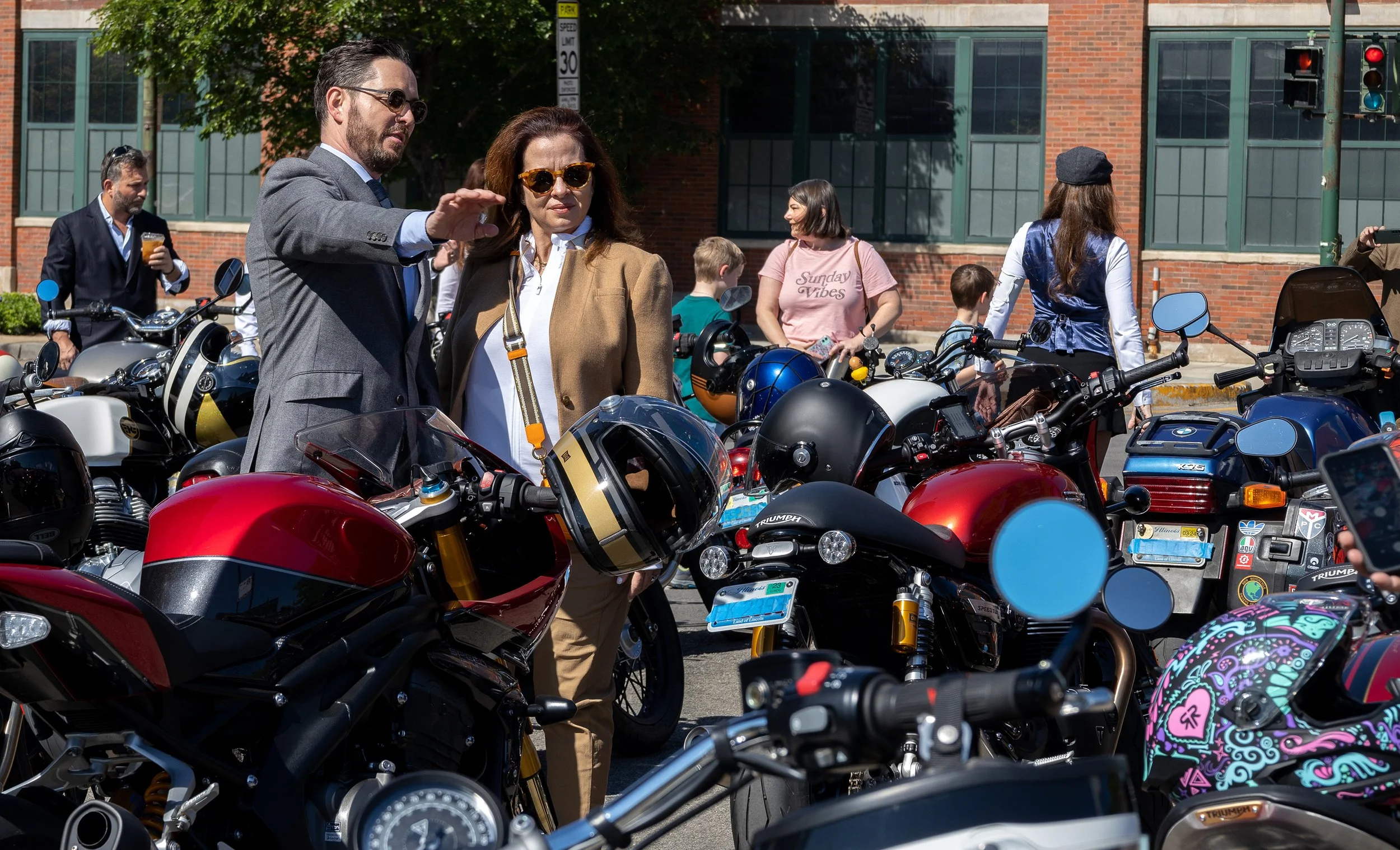 People gathered around and looking at motorcycles parked on the street, with a brick building and traffic light in the background.
