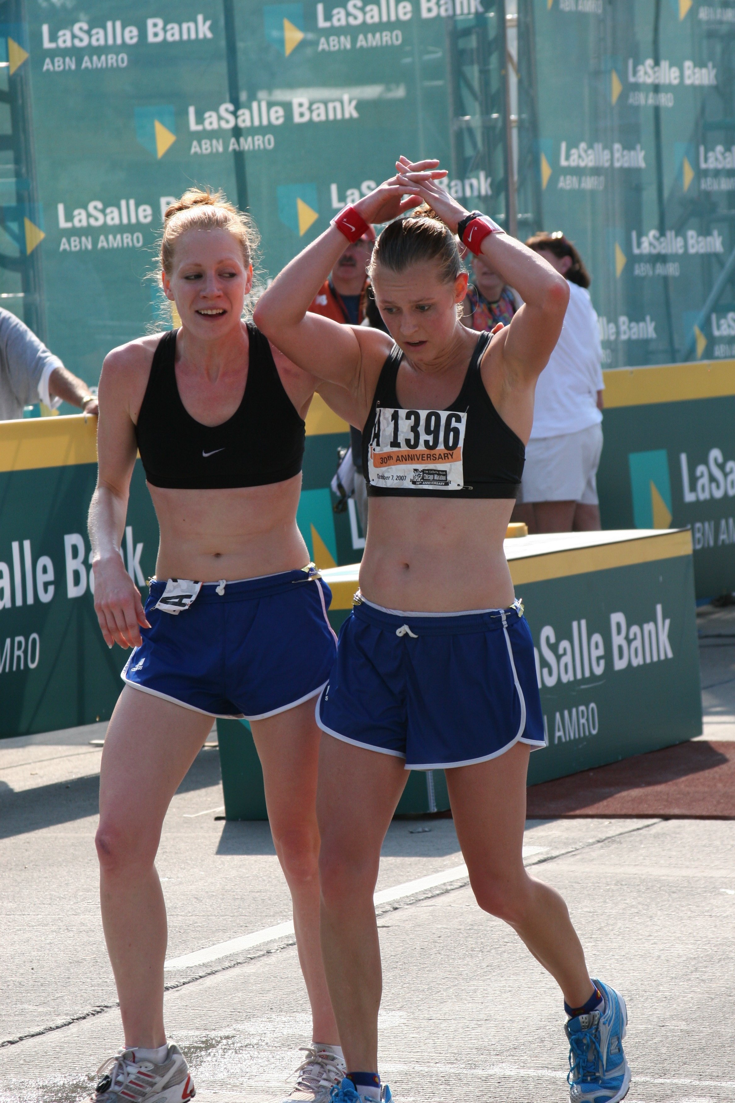 Two female marathon runners, wearing black sports bras and blue shorts, crossing the finish line with one supporting the other. The background features a green banner with the text 'LaSalle Bank ABN AMRO' and a few onlookers.
