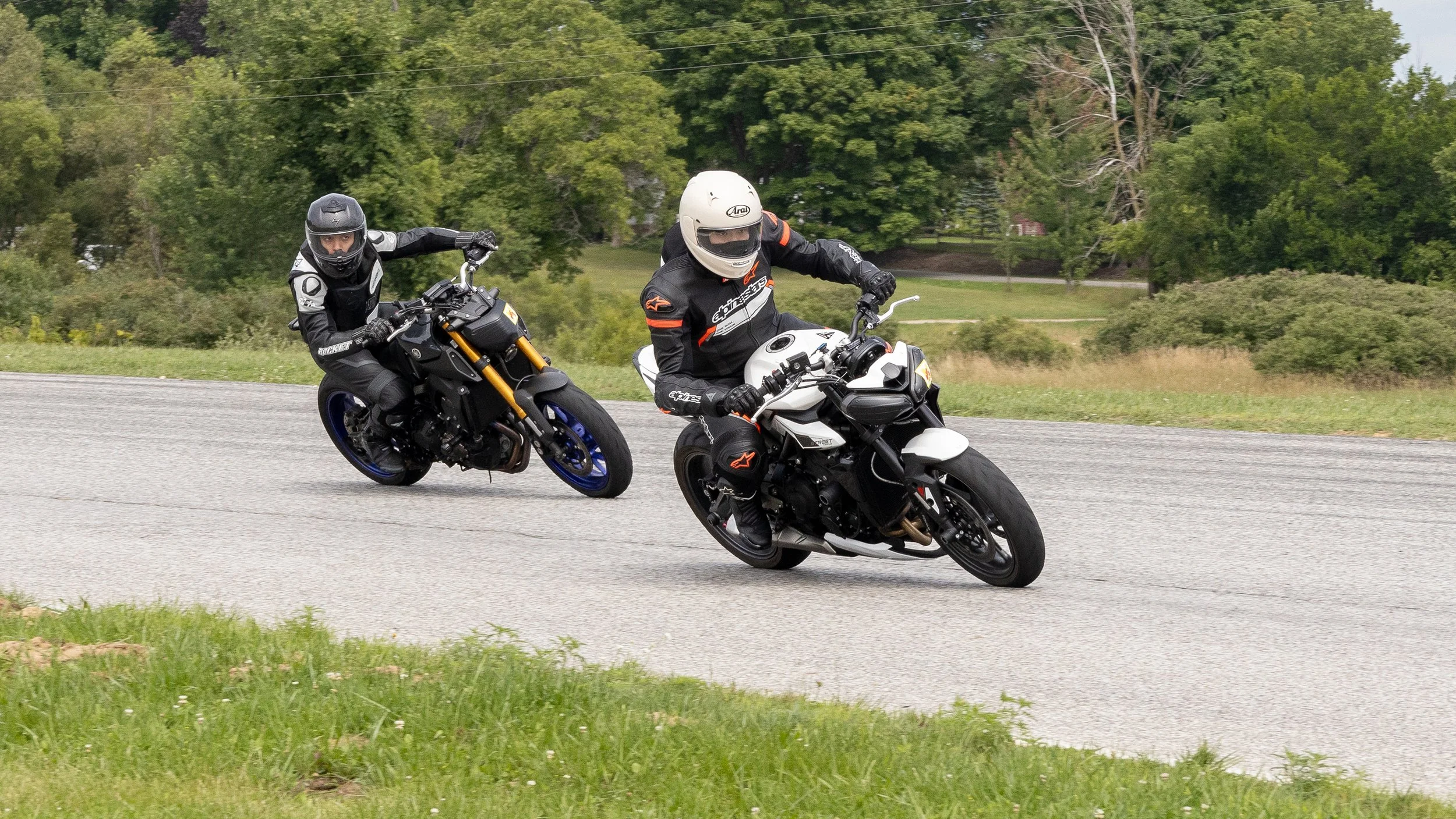 Two motorcyclists riding on a paved road through a green, tree-lined area, wearing helmets and protective gear.