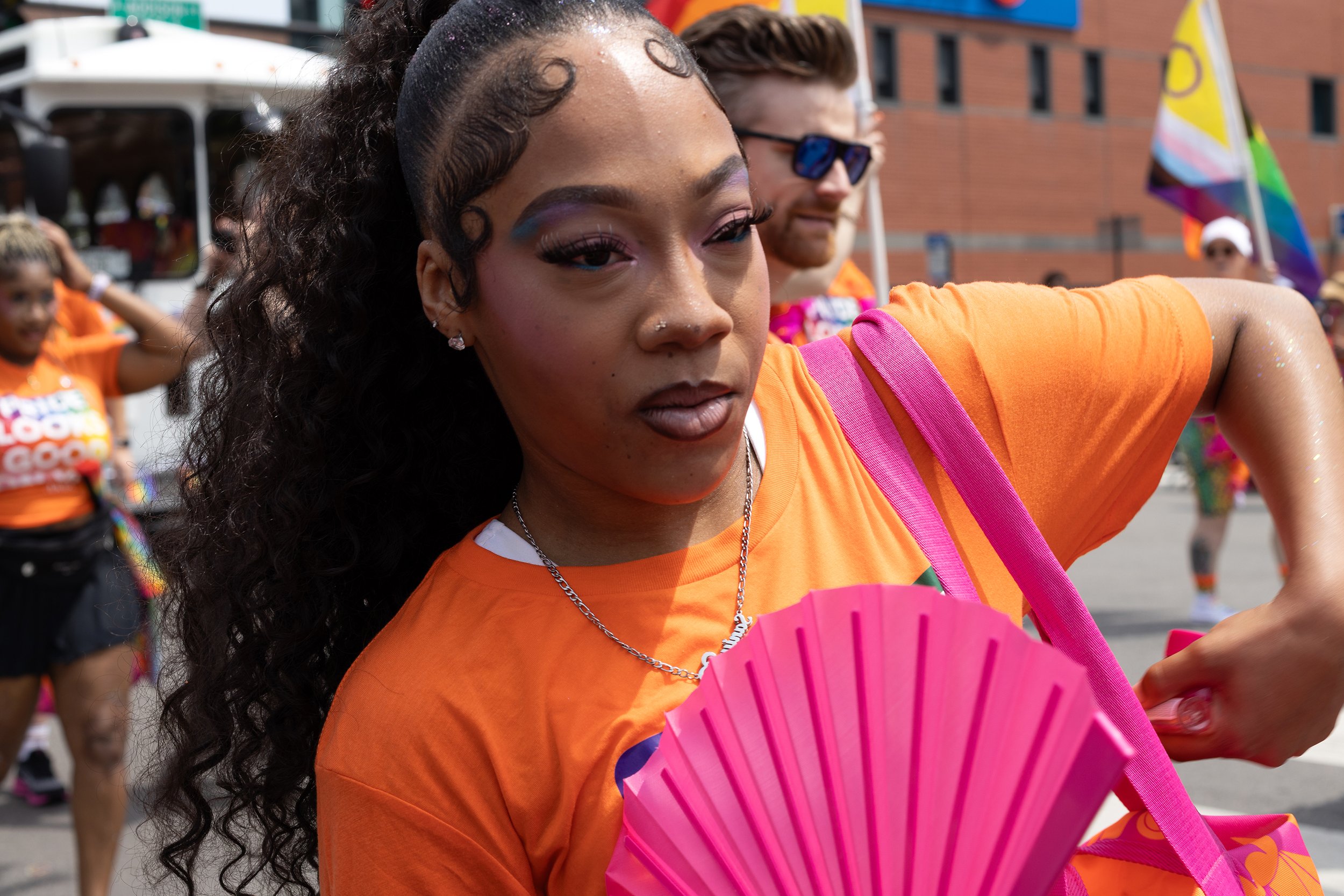 Young woman with curly black hair, colorful makeup, and jewelry holding a pink fan at a pride parade, with rainbow flags and participants in the background.