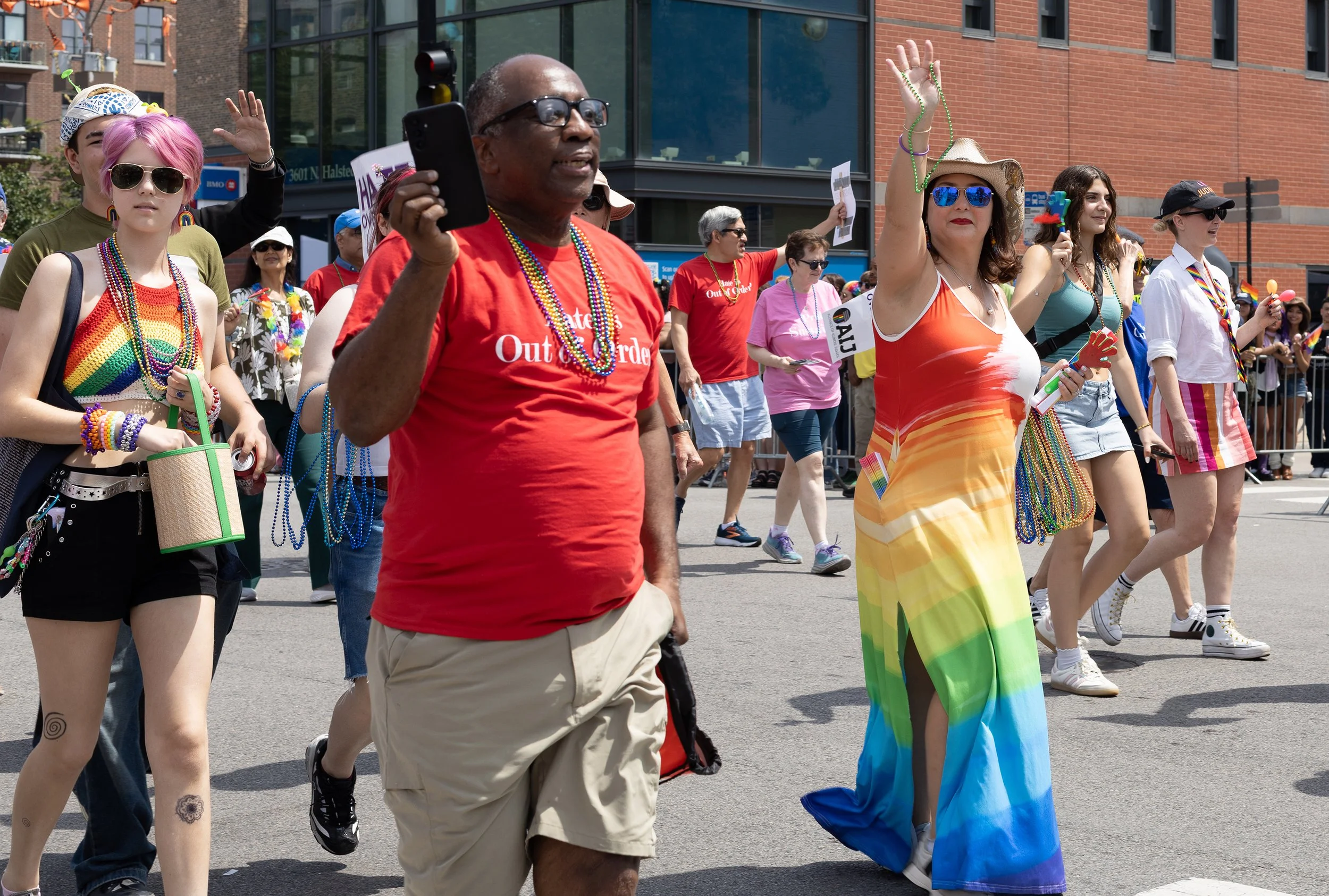 A diverse group of people participating in a pride parade, dressed in colorful clothing and accessories such as rainbow flags, beaded necklaces, and sunglasses, walking outdoors on a city street.
