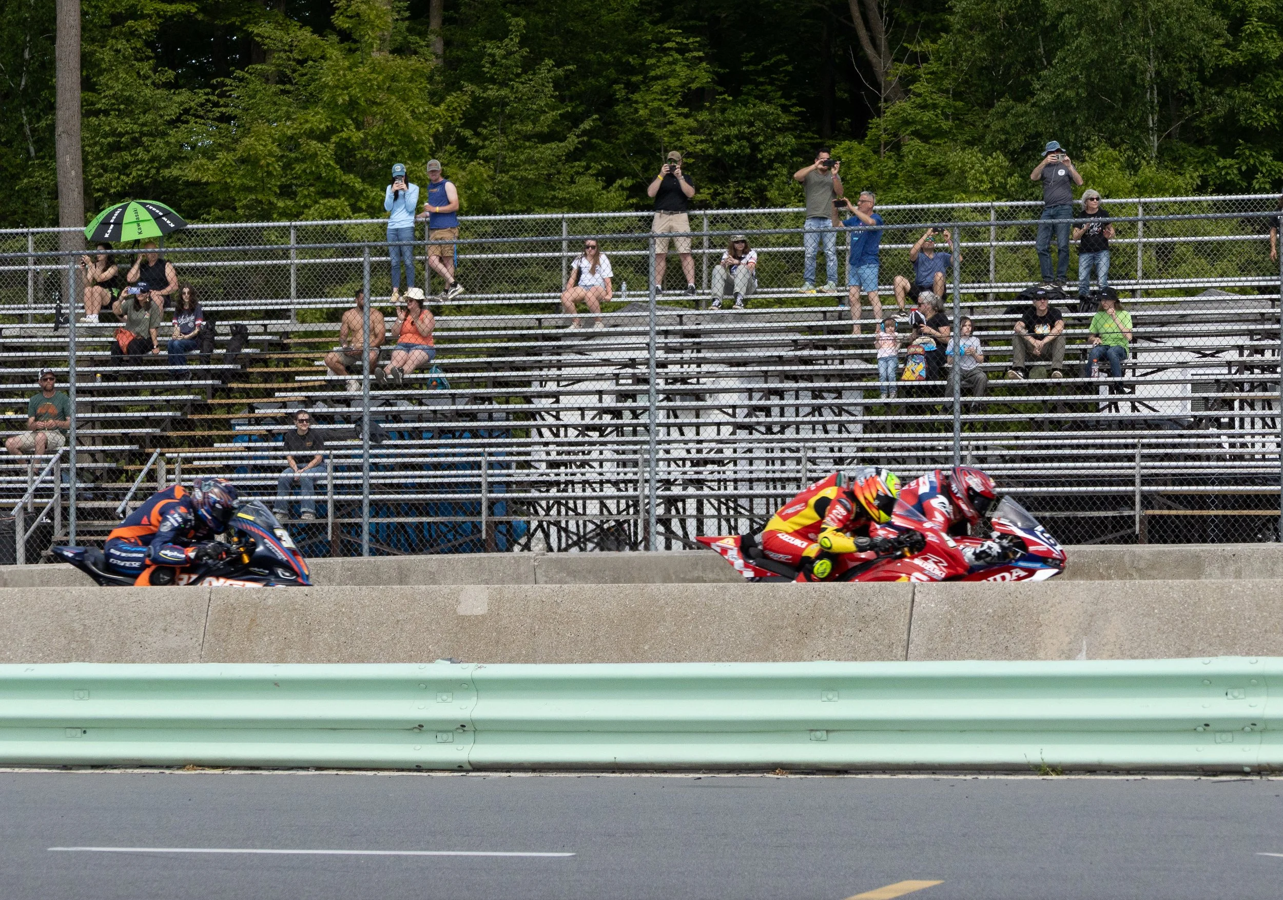 Two motorcycle racers in racing gear speeding on a racetrack, with a barrier in the foreground.