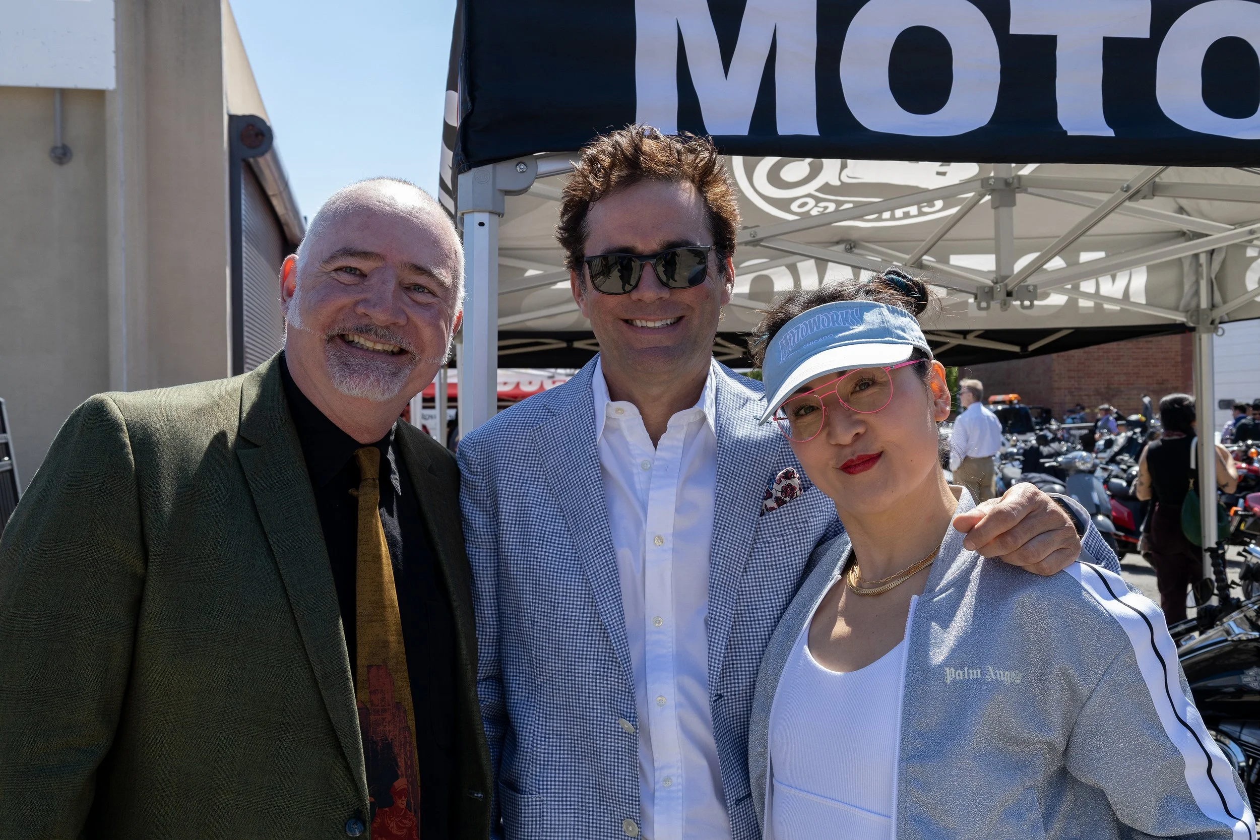 Three people posing for a photo at an outdoor event, standing under a canopy with the word 'MOTO' visible on it. People and motorcycles in the background.
