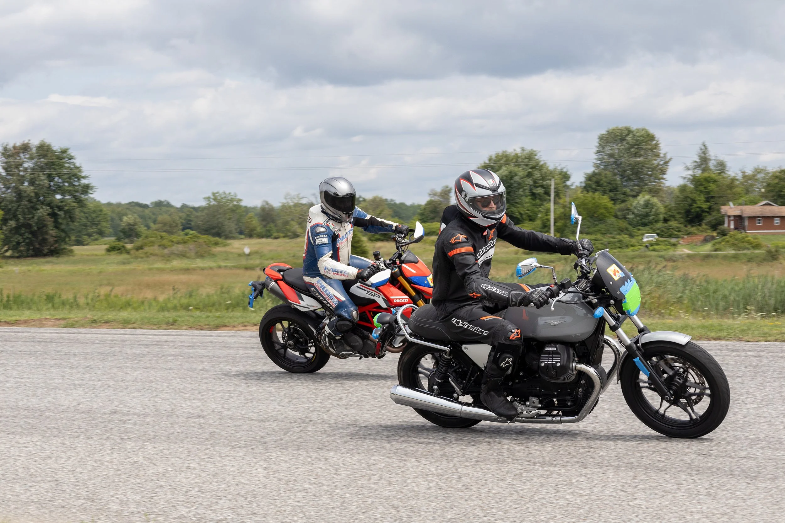 Two motorcyclists riding on a highway surrounded by greenery, wearing helmets and protective gear, with cloudy skies overhead.