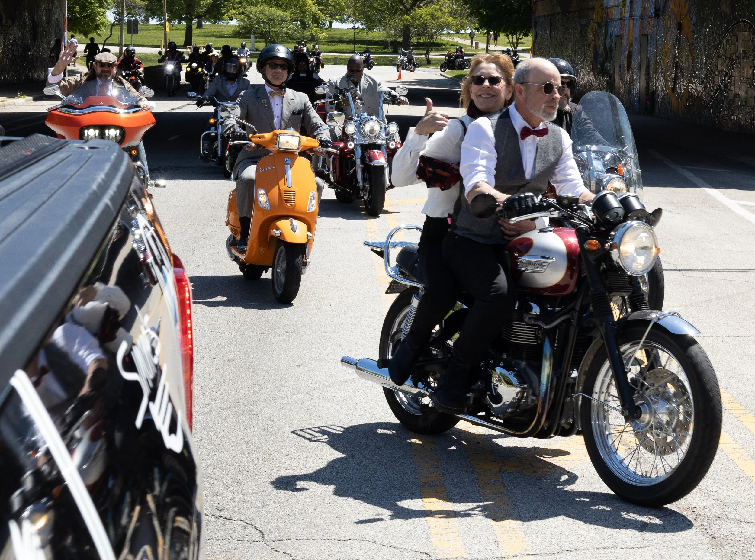 Group of people riding motorcycles and scooters outdoors on a sunny day, with some waving and smiling.