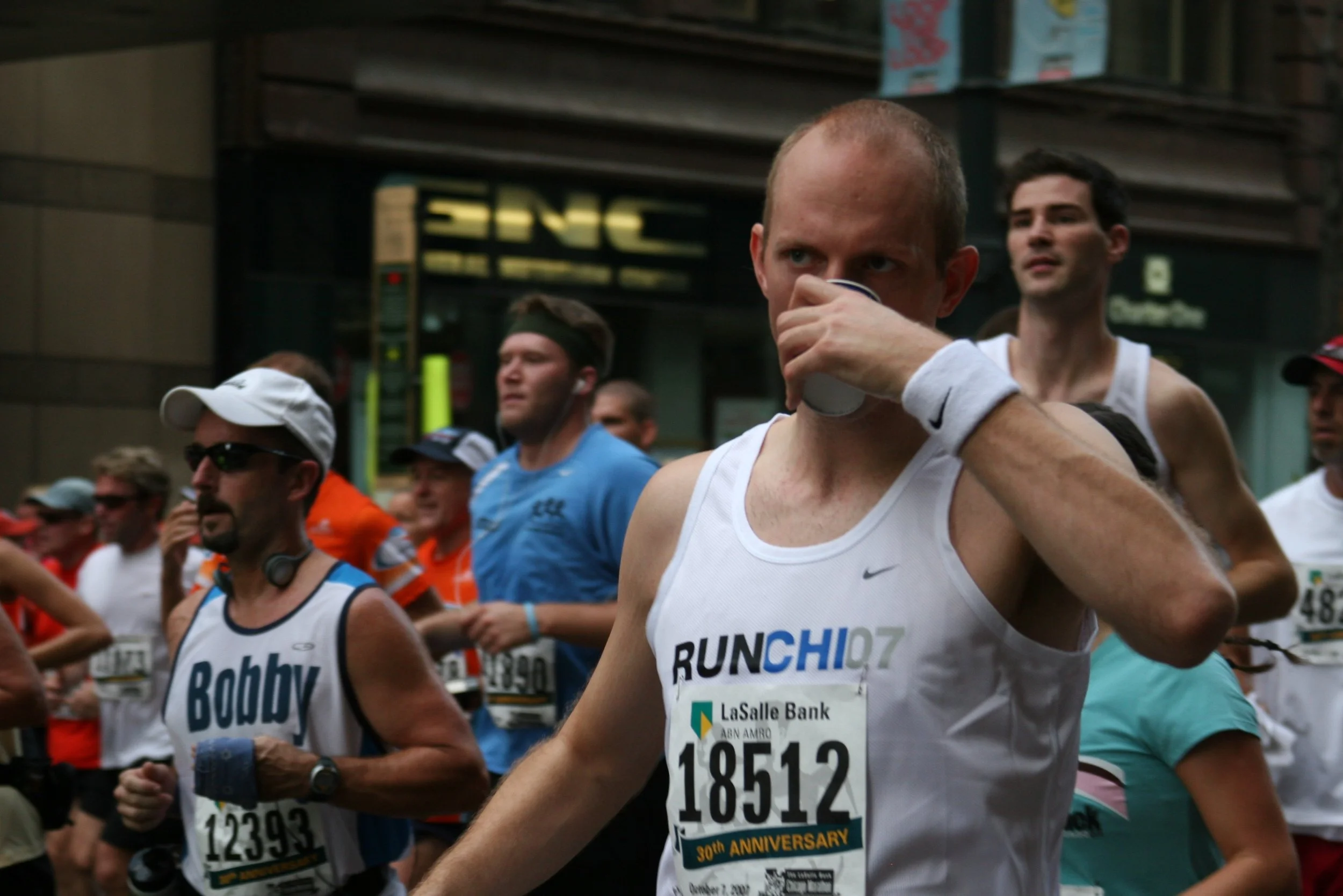 A group of runners participating in a marathon, wearing athletic clothing and race bibs, with a background showing a city street and an electronic sign.