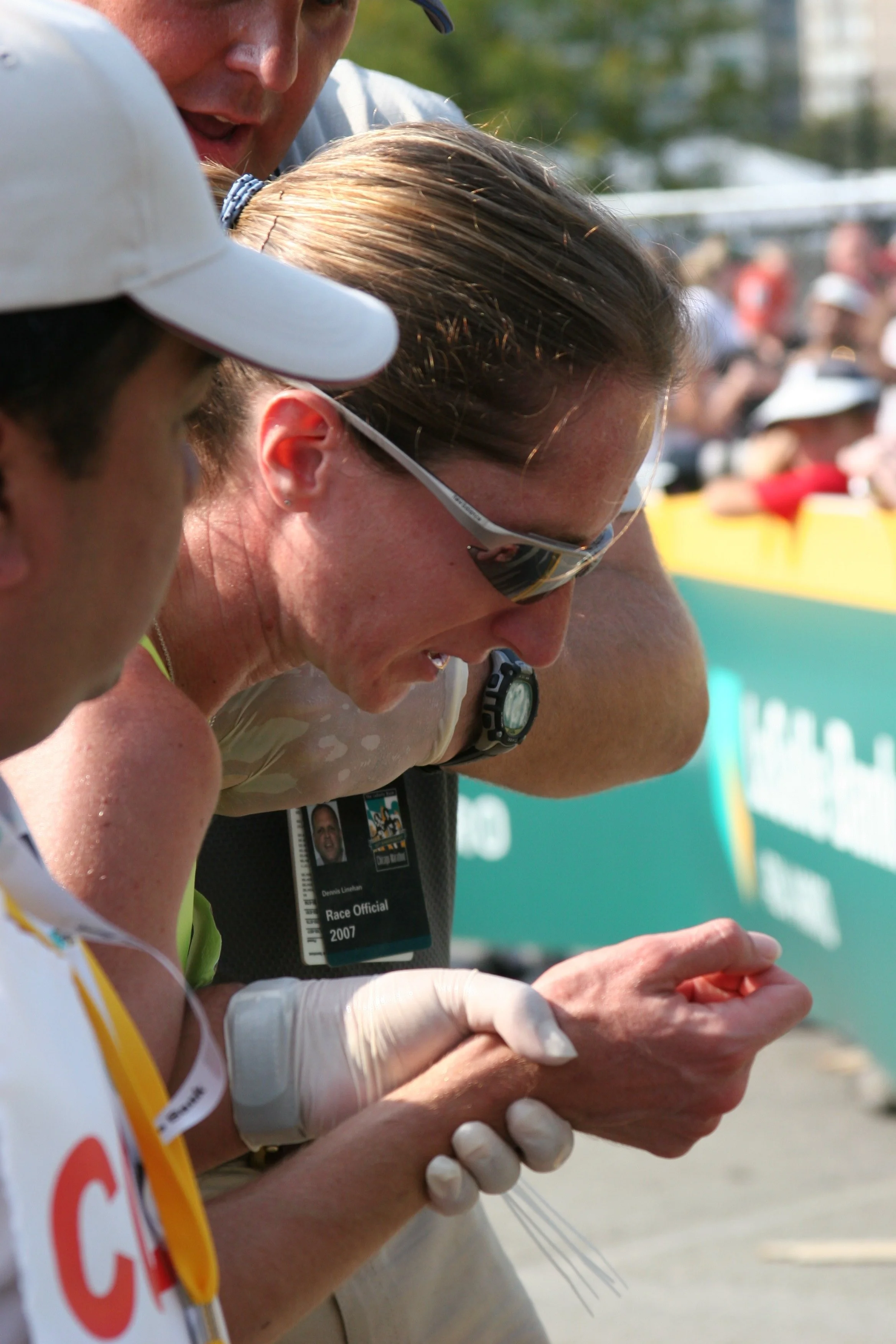 A close-up of a group of runners at a race, with some wearing sunglasses and race bibs, standing outdoors on a sunny day, with spectators and trees in the background.